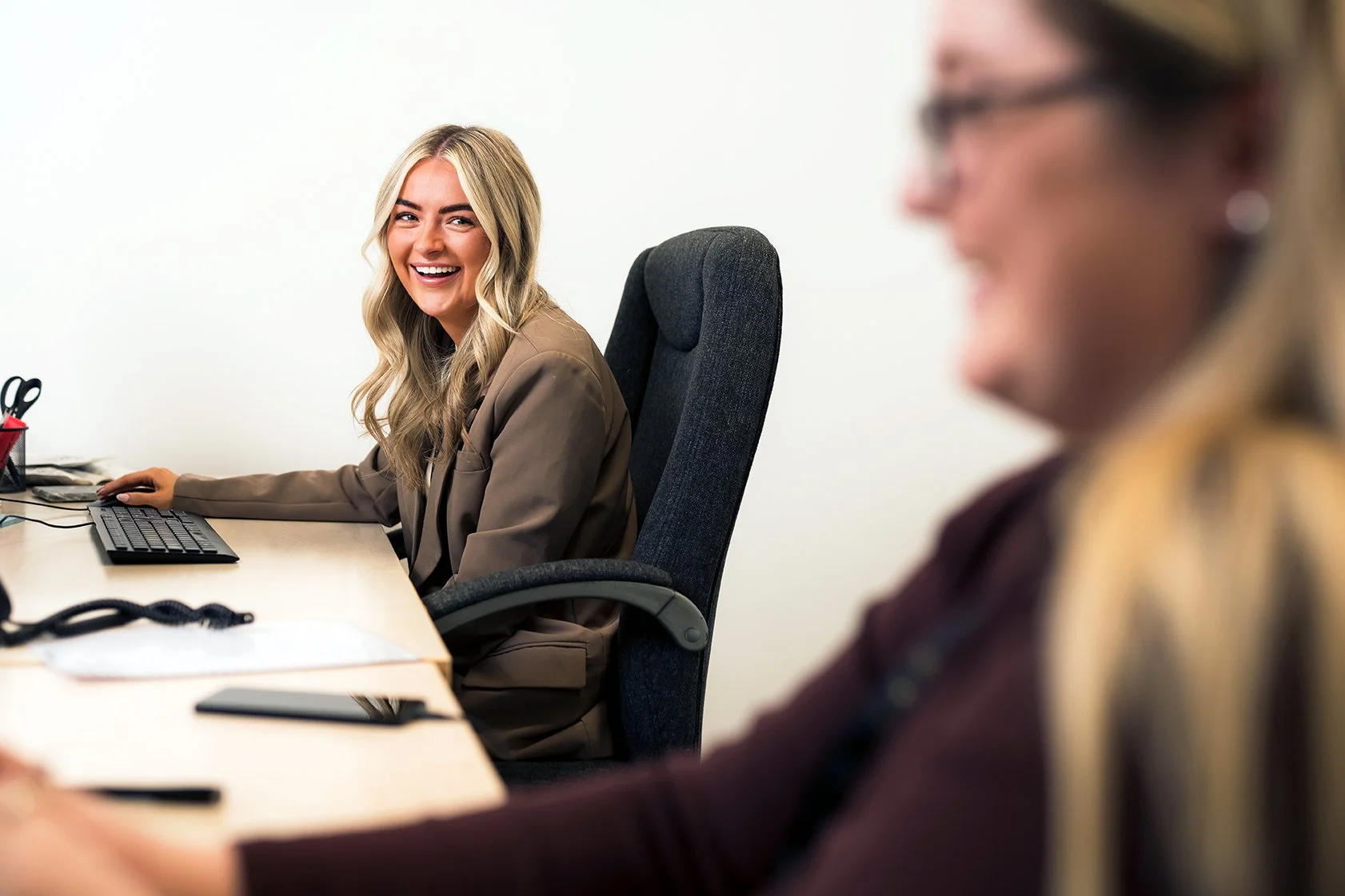 Smiling woman in a business suit sitting at a desk with a computer keyboard, in an office setting, with another woman blurred in the foreground.