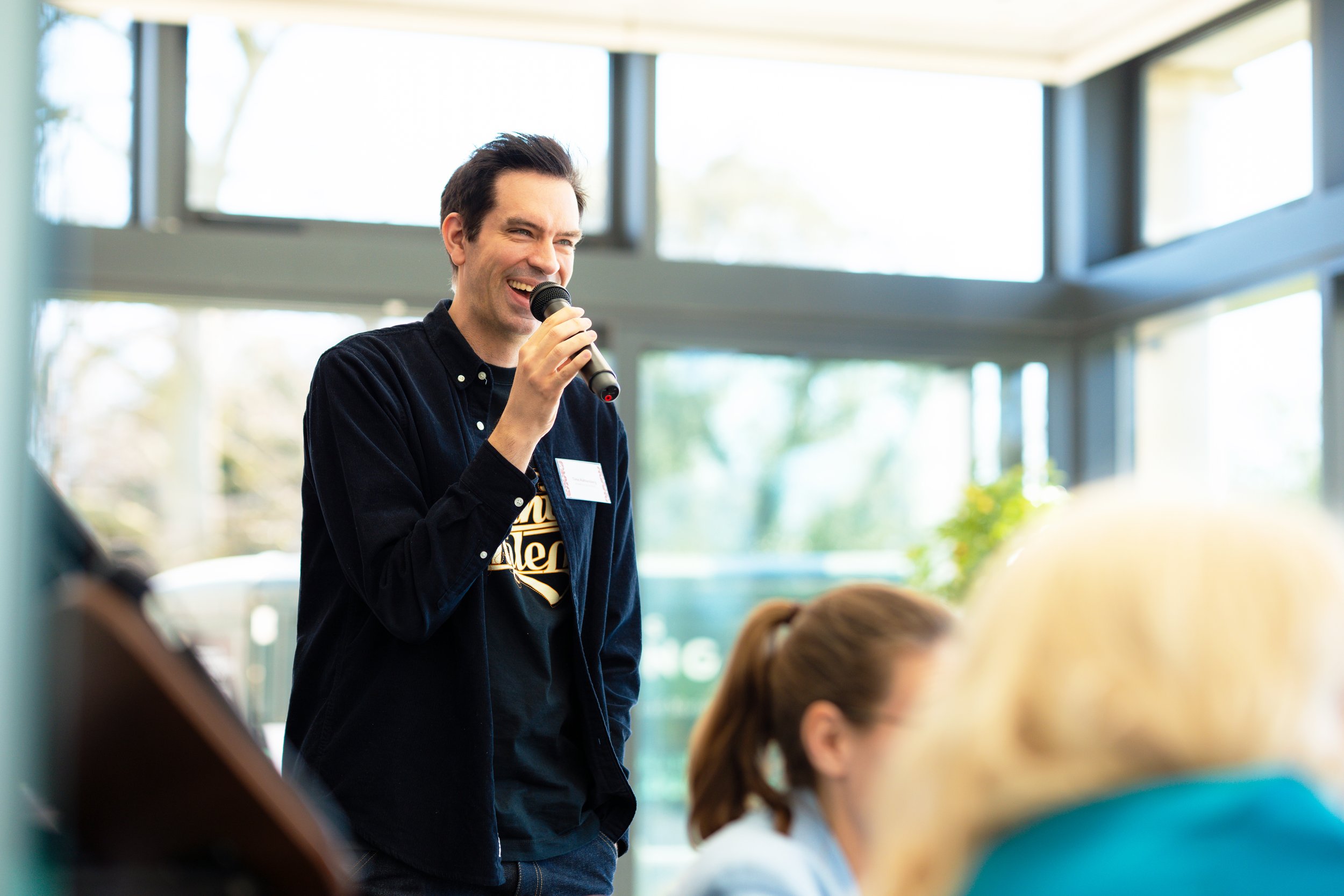 A man smiling and speaking into a microphone during a presentation or speech at indoors with large windows in the background, while people are seated in front of him.