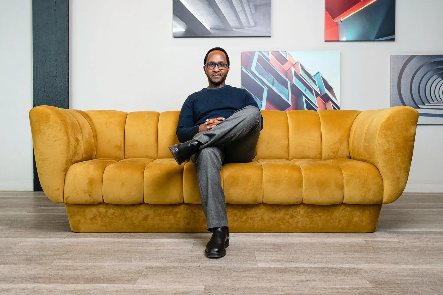 A man sitting on a yellow couch in an art gallery with abstract artwork on the wall behind him.