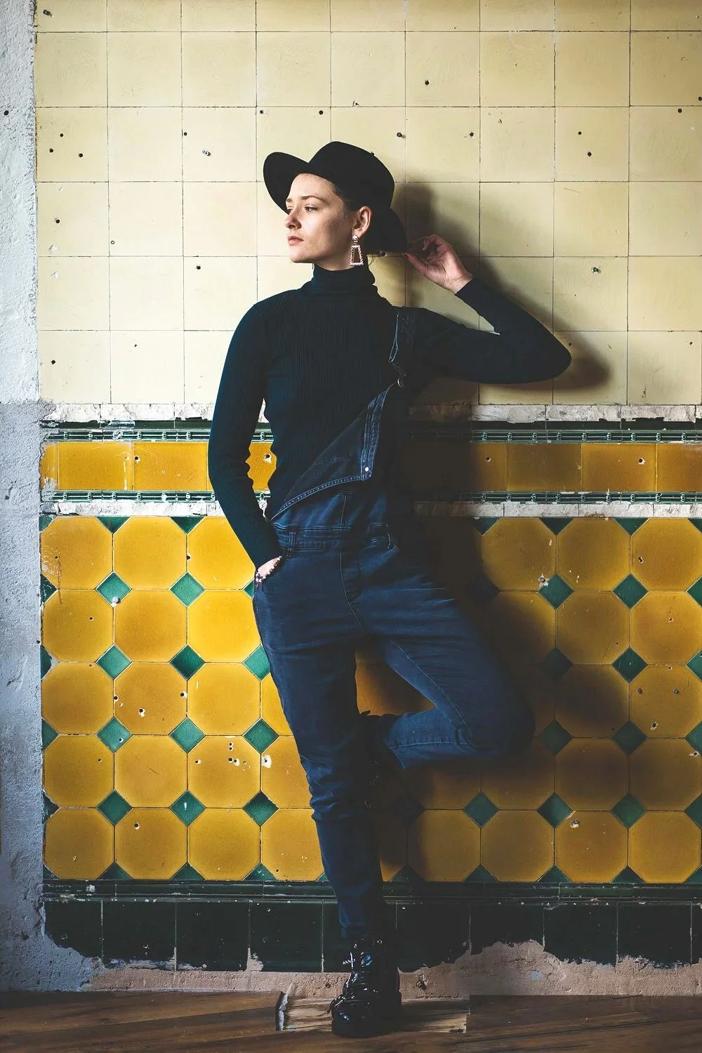 A woman in black clothing and a black hat standing against a yellow tiled wall with green accents, posing with one hand on her hat and the other in her pocket.
