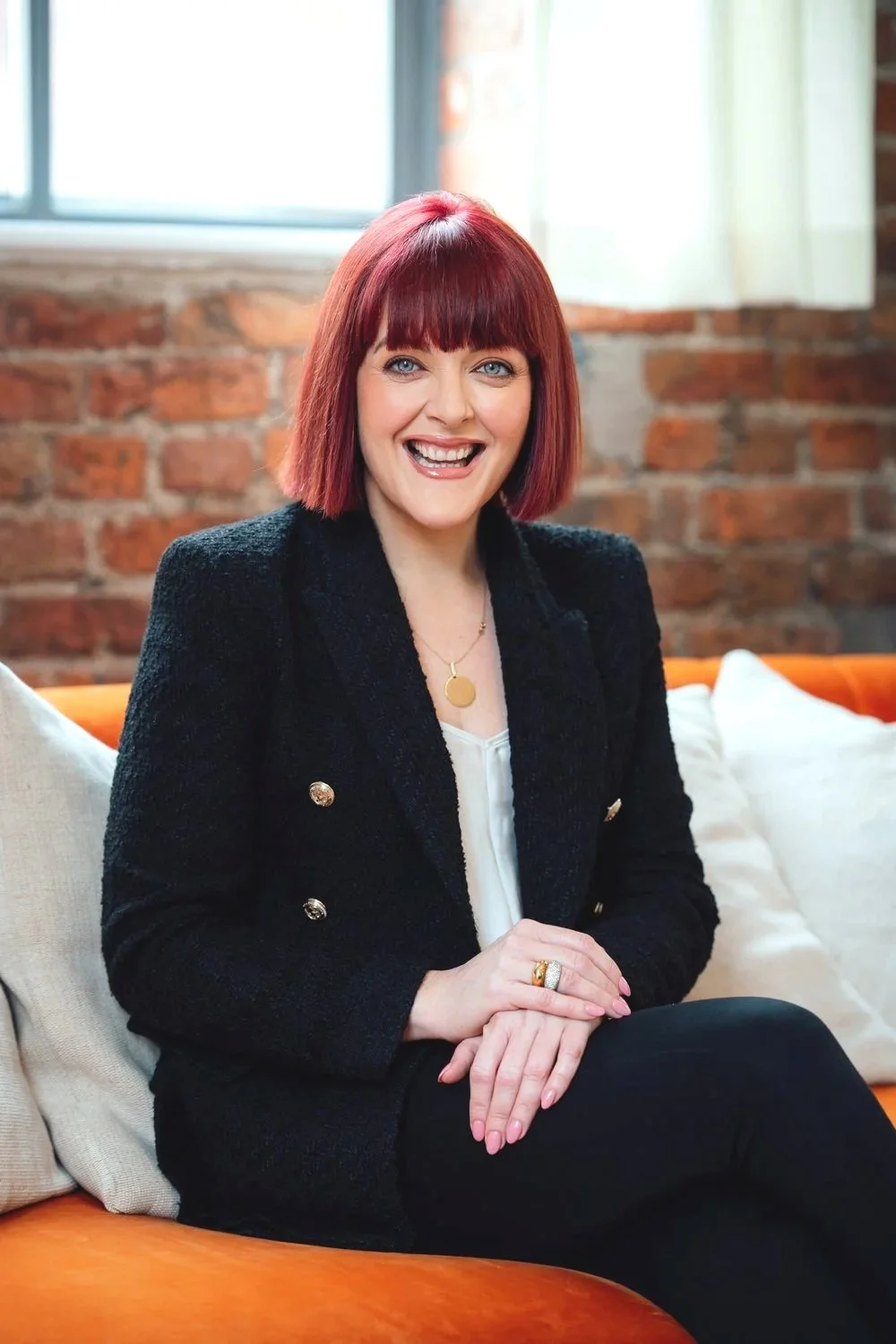 A woman with red hair, blue eyes, and a bright smile, wearing a black jacket with gold buttons and a gold pendant necklace, sitting on an orange couch with white pillows against an exposed brick wall and a window in the background.