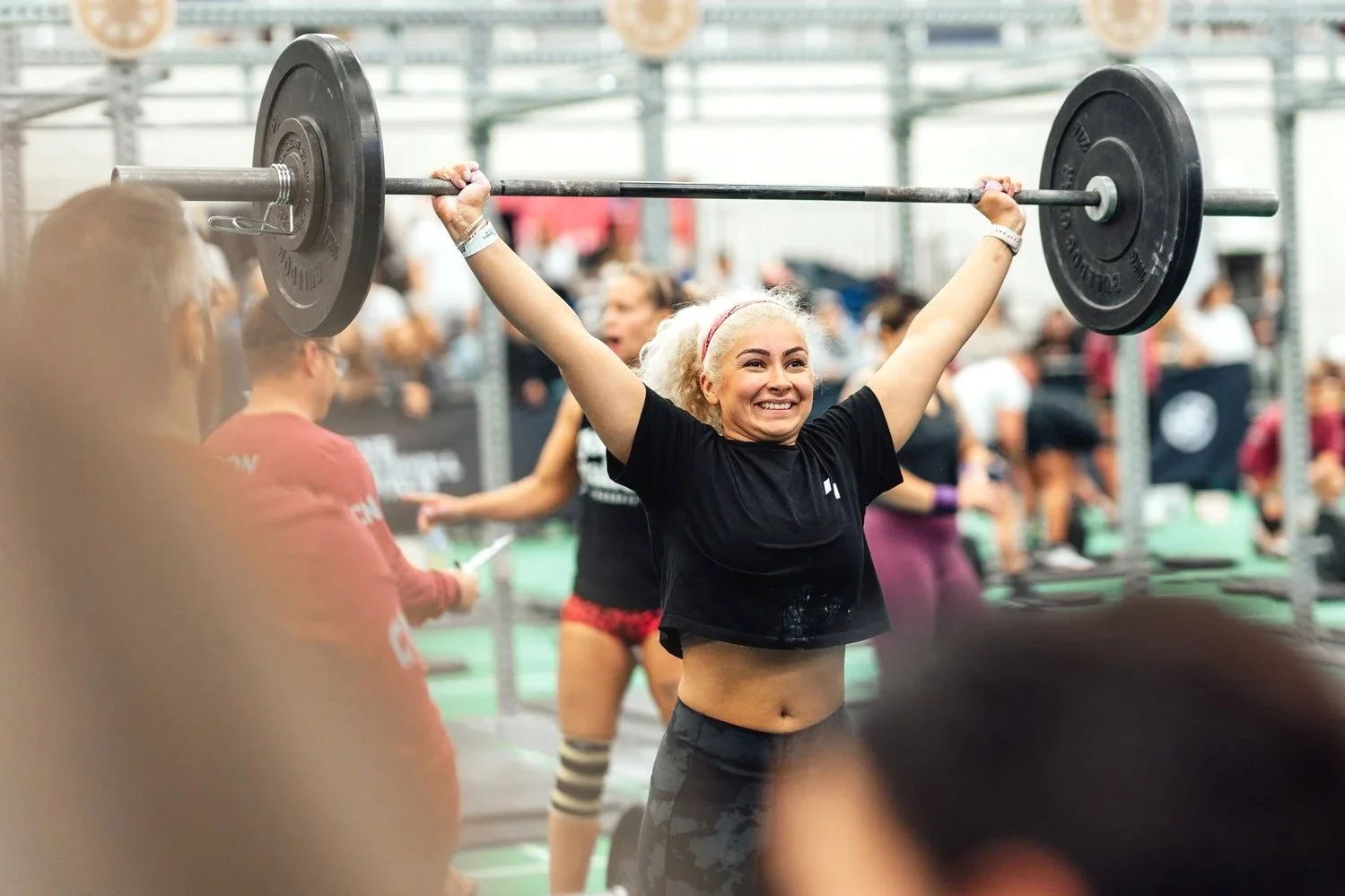 Woman with blonde curly hair lifting a barbell overhead at a fitness competition with a joyful expression.