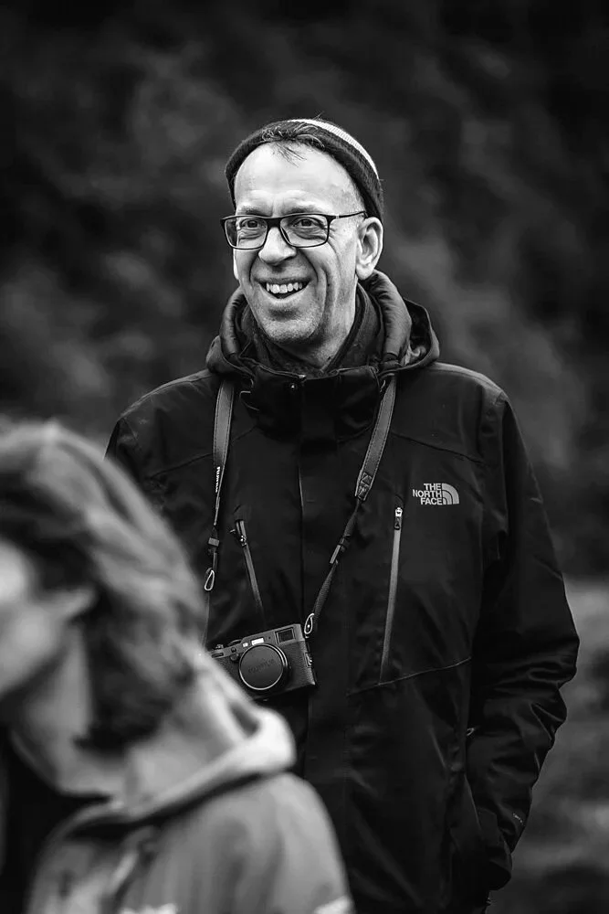 Man with glasses, beanie, and a North Face jacket smiling outdoors with a camera hanging around his neck.