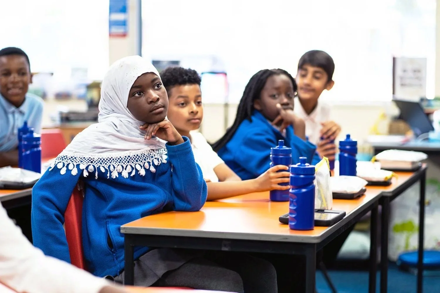 School children sitting at desks listening attentively, with water bottles and school supplies in a classroom.