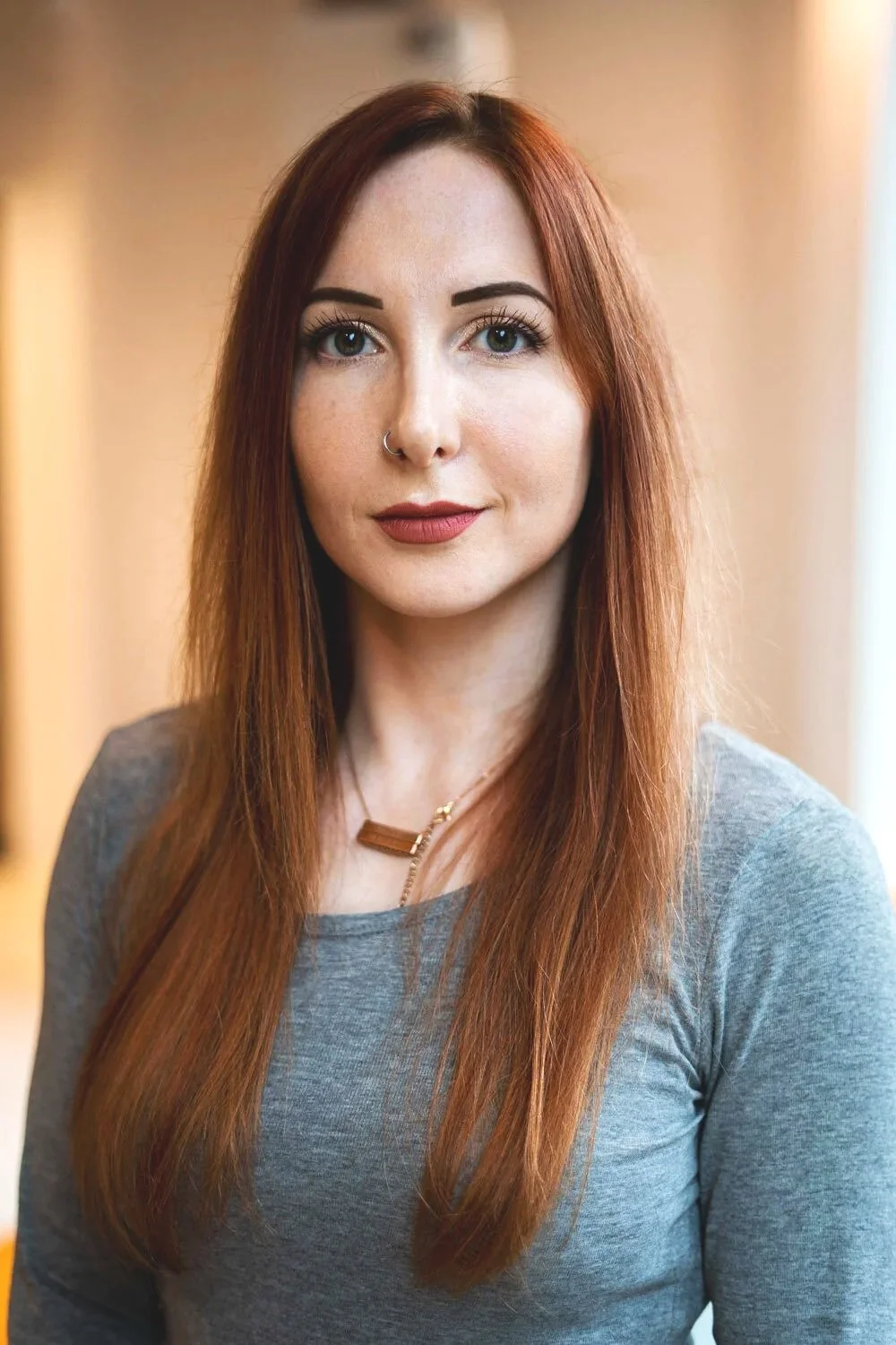 A young woman with long red hair, wearing a gray shirt and a necklace, looking directly at the camera with a soft smile.
