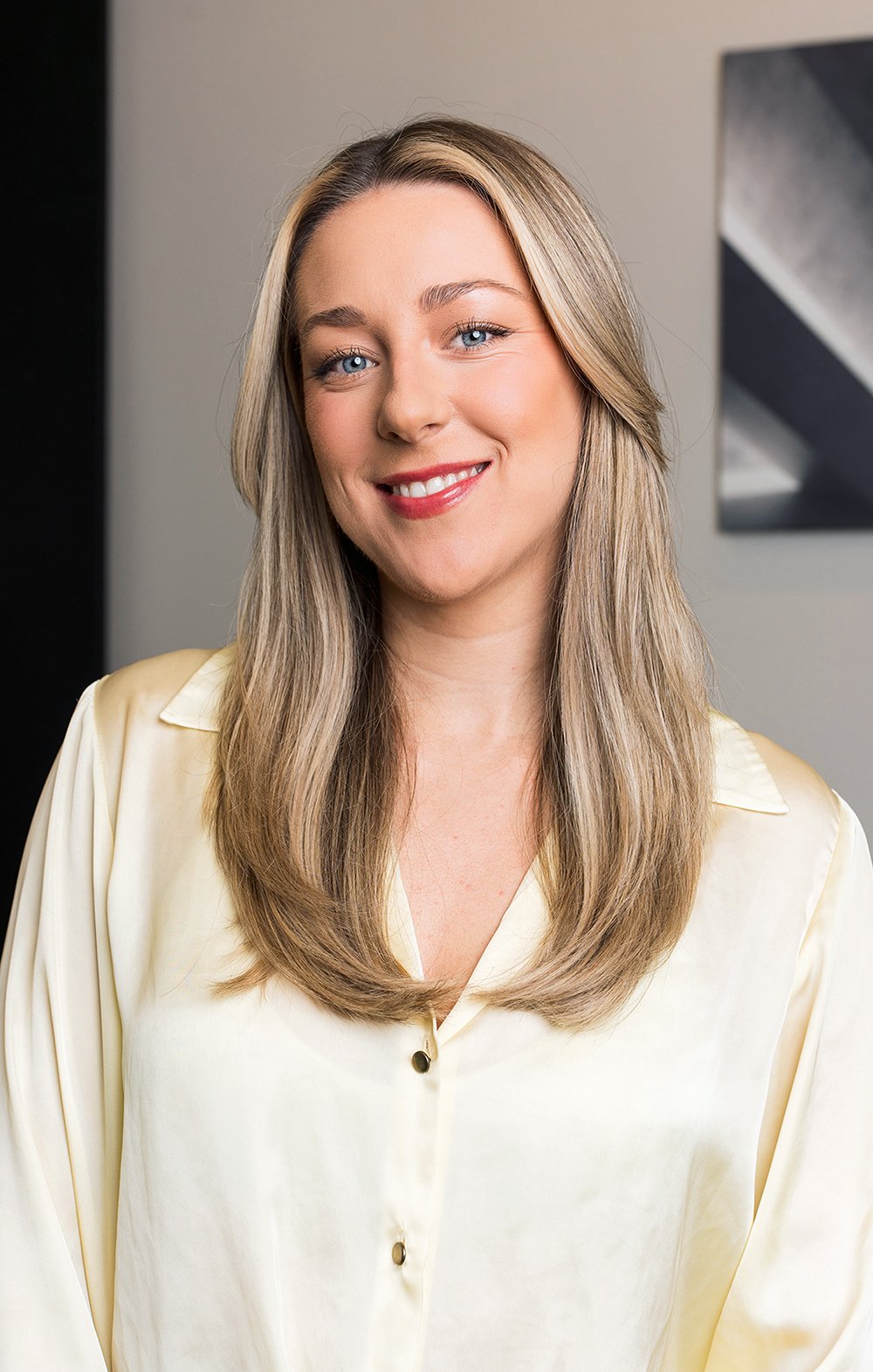 A woman with long blonde hair and blue eyes smiling, wearing a light-colored blouse, standing indoors with a neutral background.