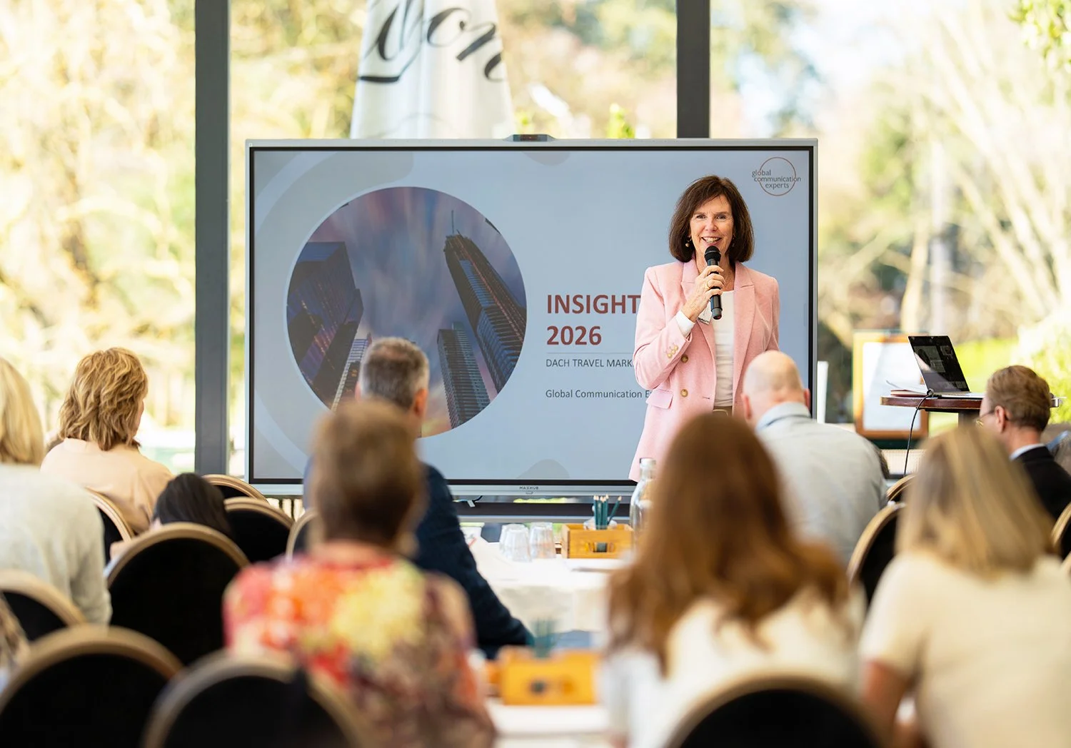 A woman in a pink blazer is speaking into a microphone in front of a large screen at a conference, with the audience seated and listening attentively. The screen displays a presentation titled "INSIGHT 2026" with an image of tall buildings.