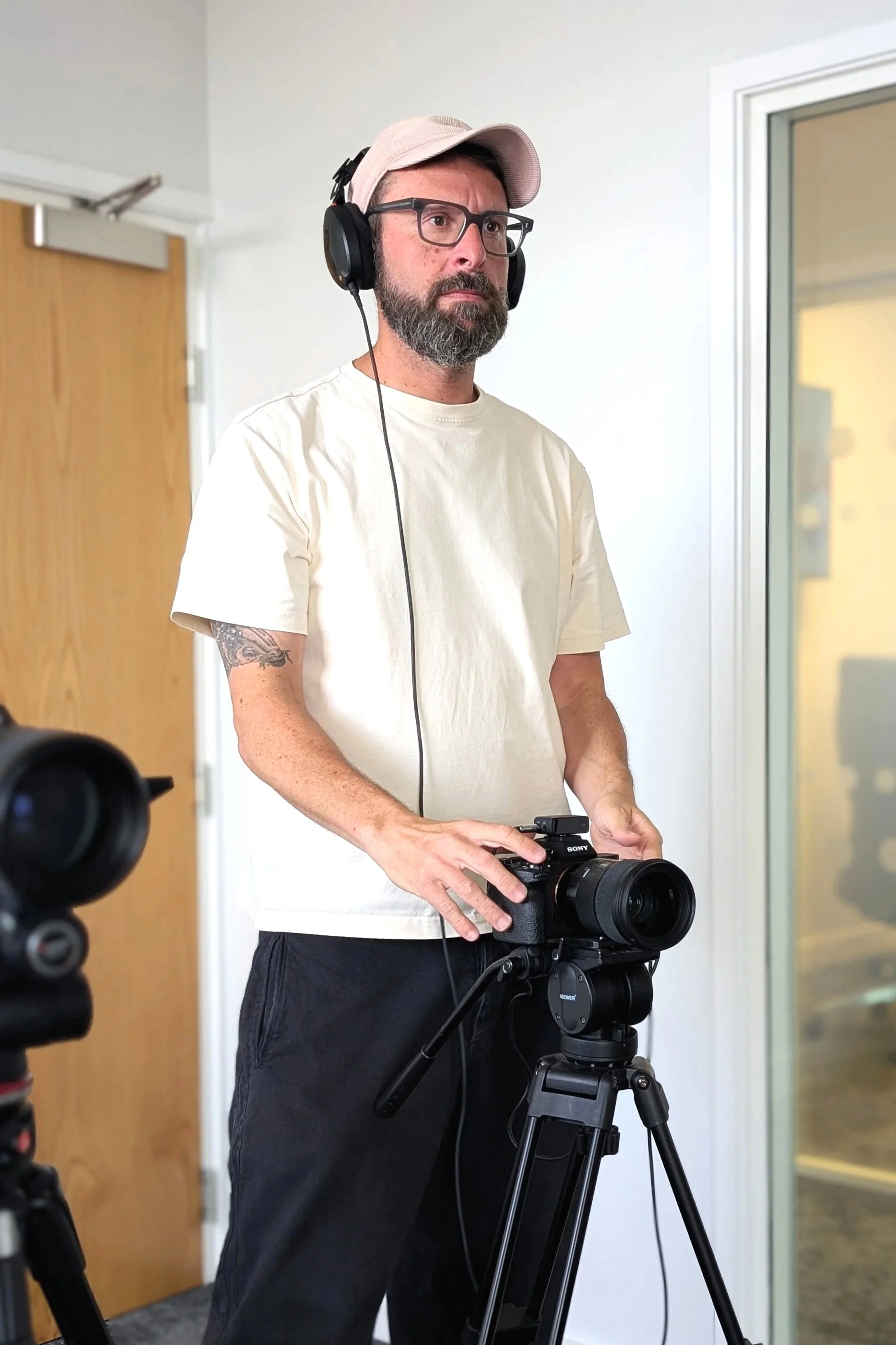 A man with glasses, a beard, and a tattooed arm wearing a pink cap and headphones, holding a camera on a tripod in a room with a door and glass window.