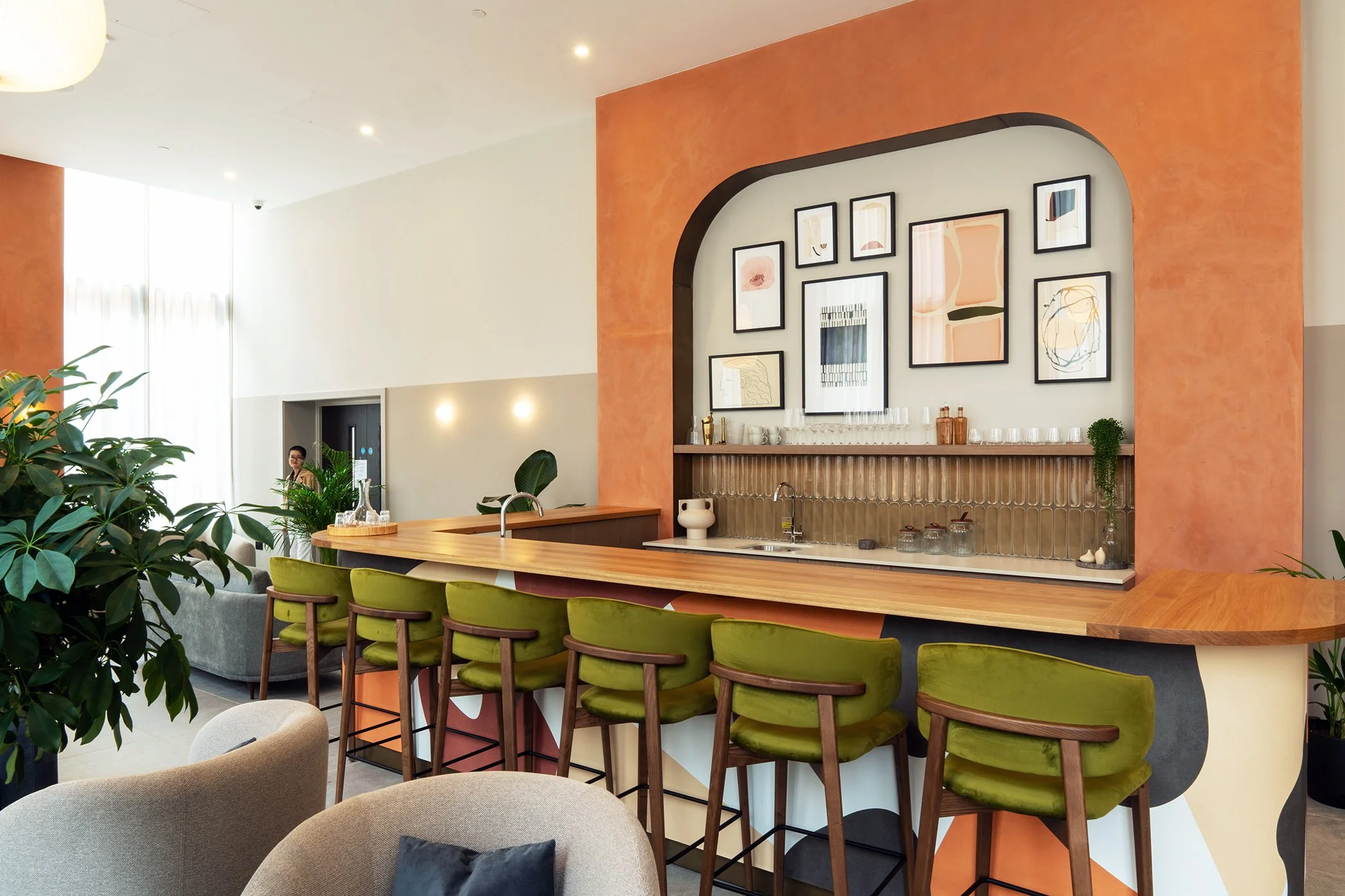 A stylish restaurant interior with a bar counter and green upholstered barstools, framed abstract artwork on the wall, and large windows allowing natural light.