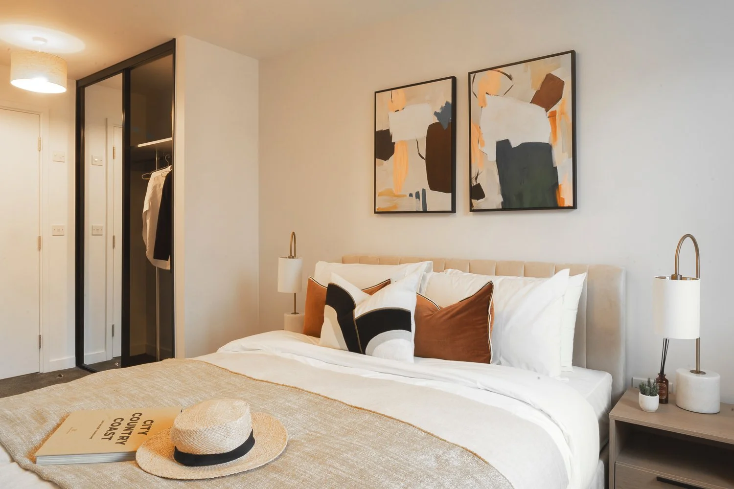 A neatly made bed with multiple decorative pillows, a straw hat on a book titled 'City Country Coast,' and a bedside table with a lamp and small decorative items in a modern bedroom with abstract artwork on the wall.