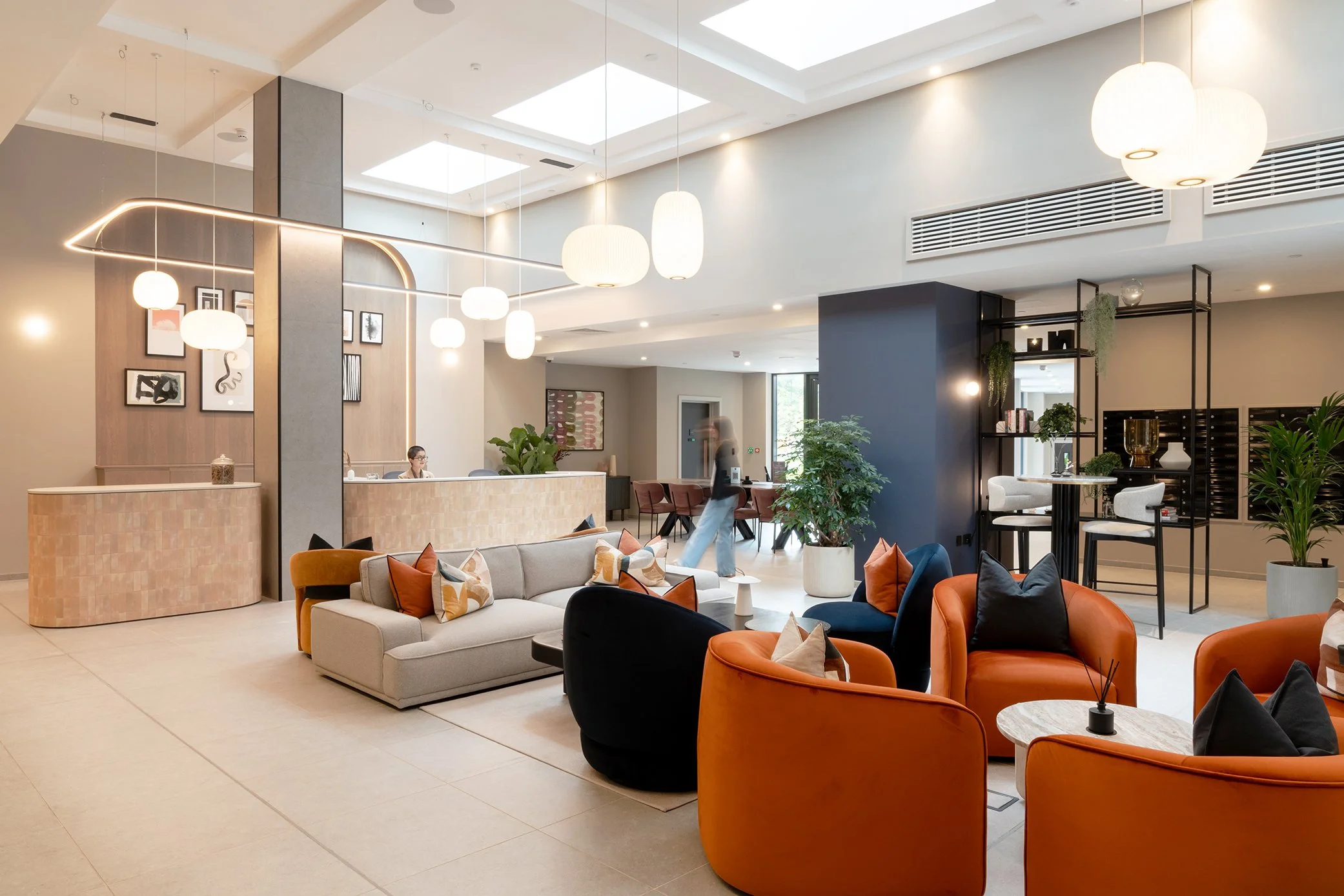 Interior of a modern hotel lobby with various seating arrangements, including orange, black, and beige chairs, a curved beige reception desk, artwork on the wall, plants, and hanging lights.