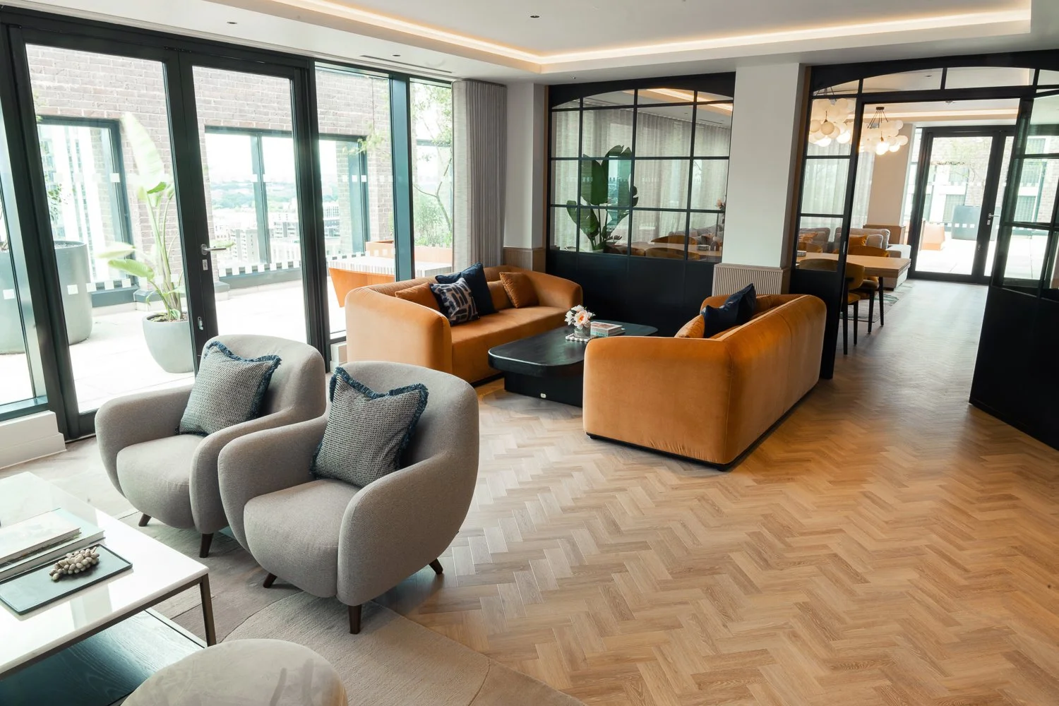Modern living room with large glass doors, tan sofas with decorative pillows, and gray armchairs with cushions, wood floors, and a balcony visible outside.