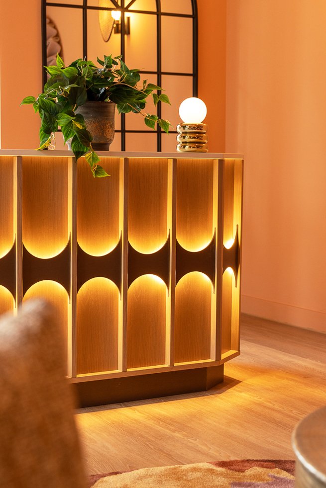 Decorative wooden cabinet with illuminated curved panels, a potted plant, a modern table lamp, and a mirror with black grid design in the background.