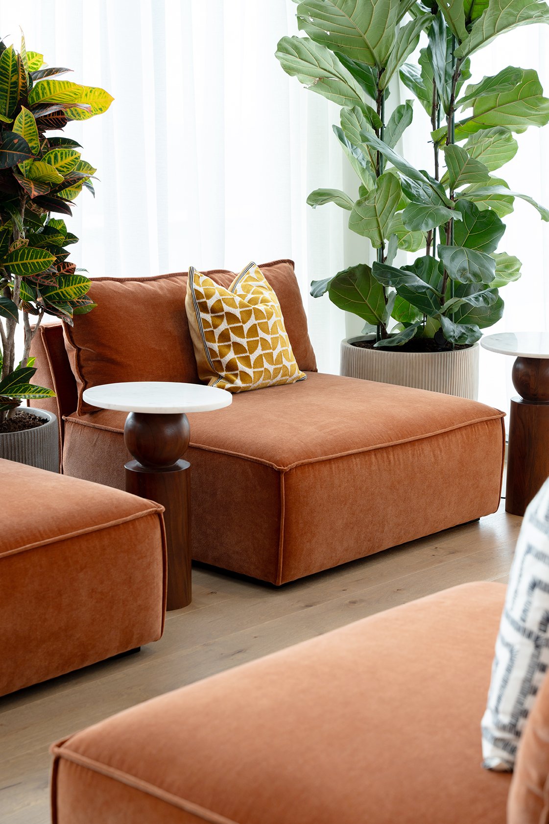 Living room with coral-colored sofa, yellow patterned throw pillow, two small side tables with white tops and dark wood bases, and large green potted plants near white curtains.