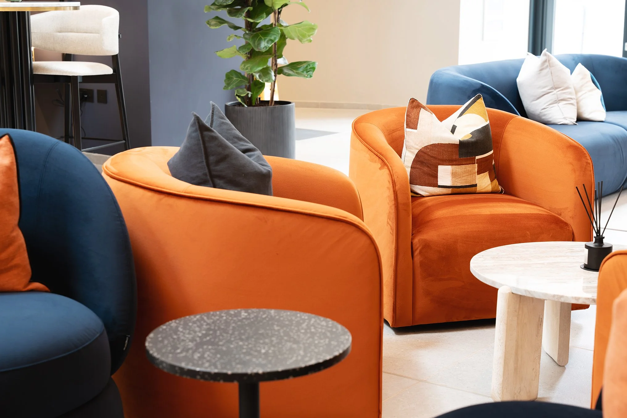 Colorful modern sitting area with orange and blue upholstered chairs, a white marble side table, a potted plant, and decorative pillows.