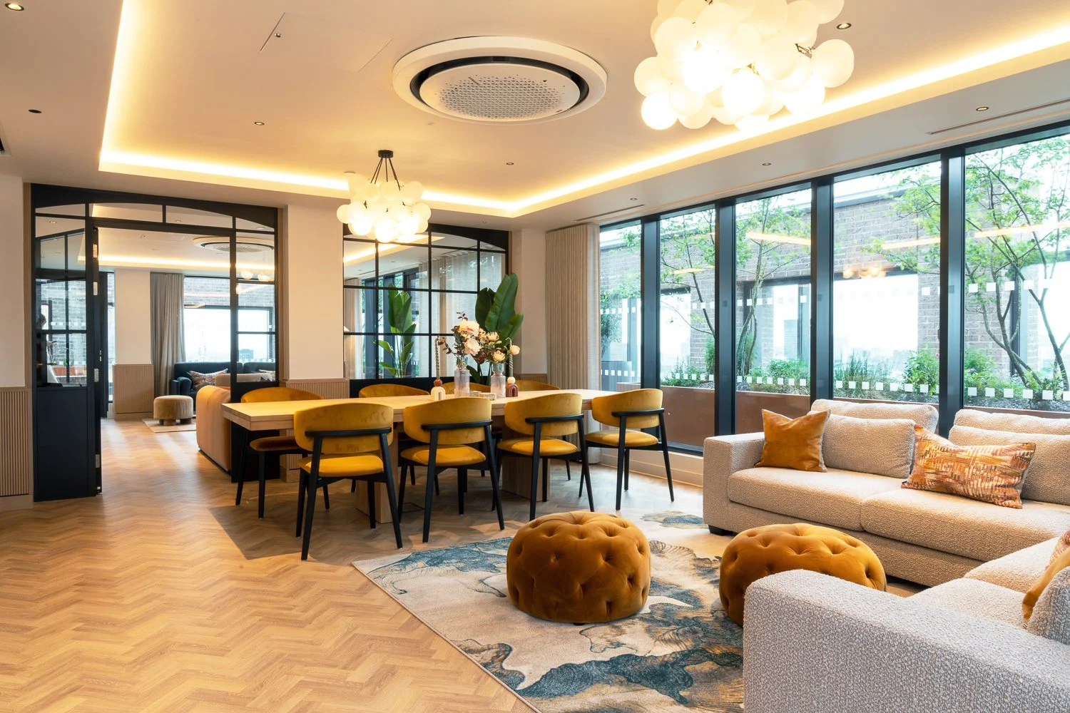 Modern living room with large windows, beige sofas, mustard-colored chairs around a wooden dining table, tufted ottomans, and a floral rug. Pendant lights and natural light brighten the space.