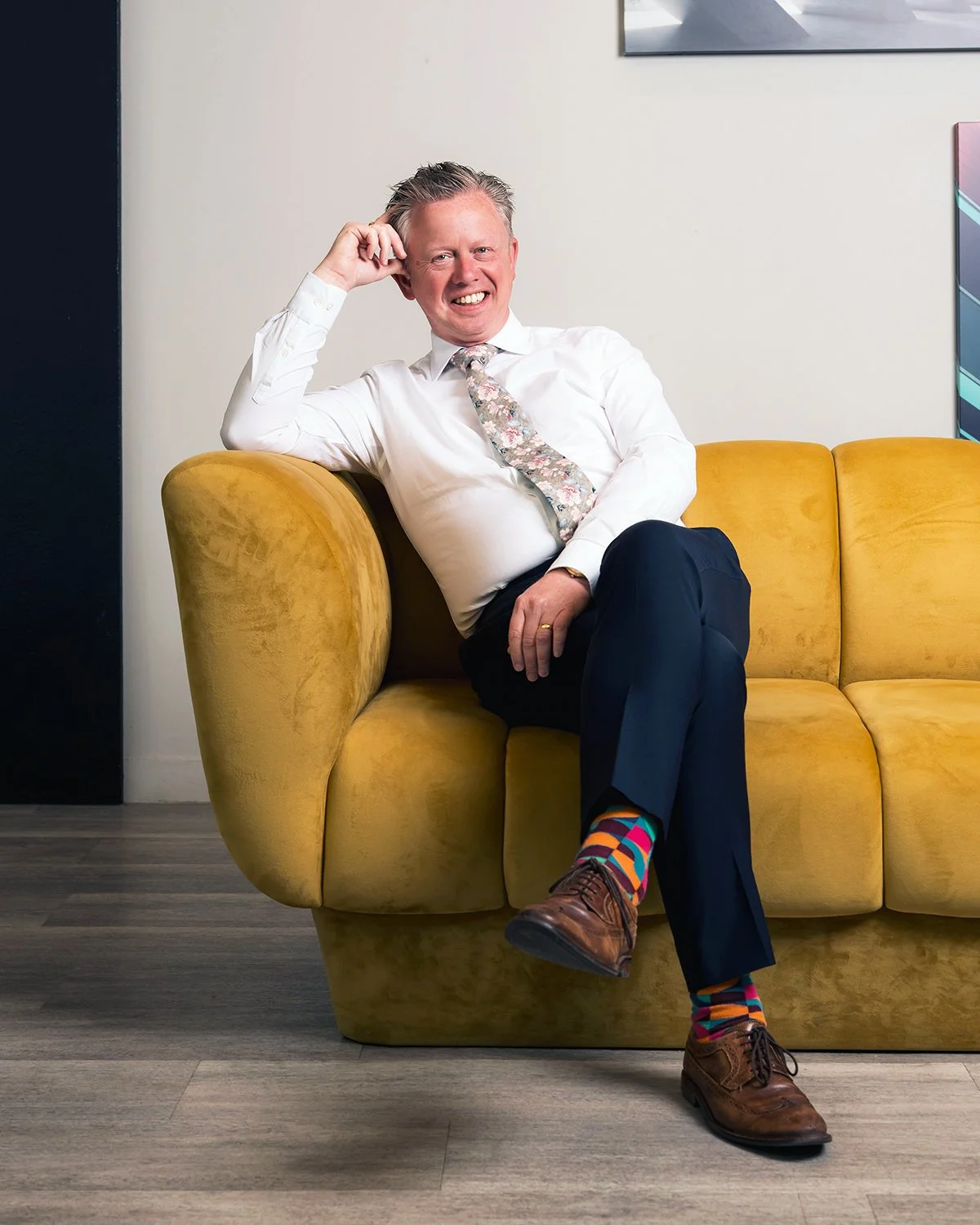 A smiling man in a white shirt and floral tie, sitting on a yellow couch with legs crossed, wearing colorful socks and brown shoes, in a modern room with artwork on the wall.