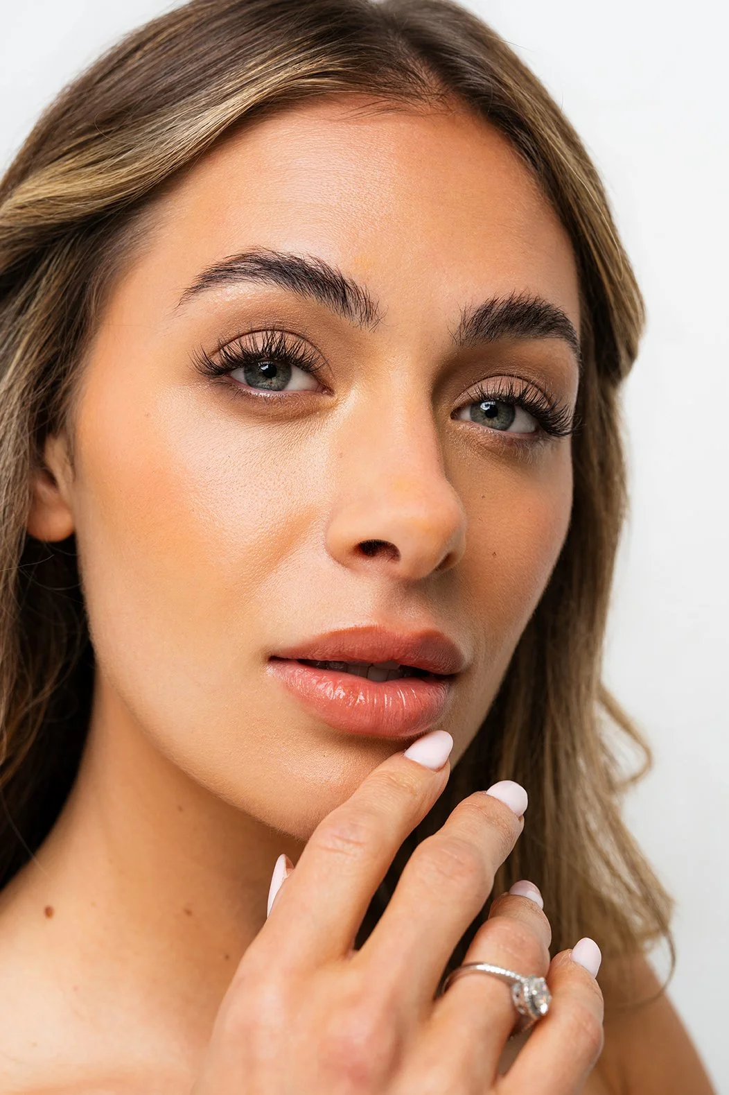 Close-up of a woman with light skin, wavy brown hair, blue eyes, and natural makeup, touching her chin with her fingers, wearing a ring.