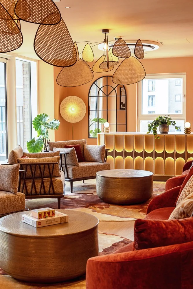 A cozy modern lounge with plush orange and beige armchairs, round coffee tables, large potted plants, and warm lighting, featuring a decorative bar with backlit panels.