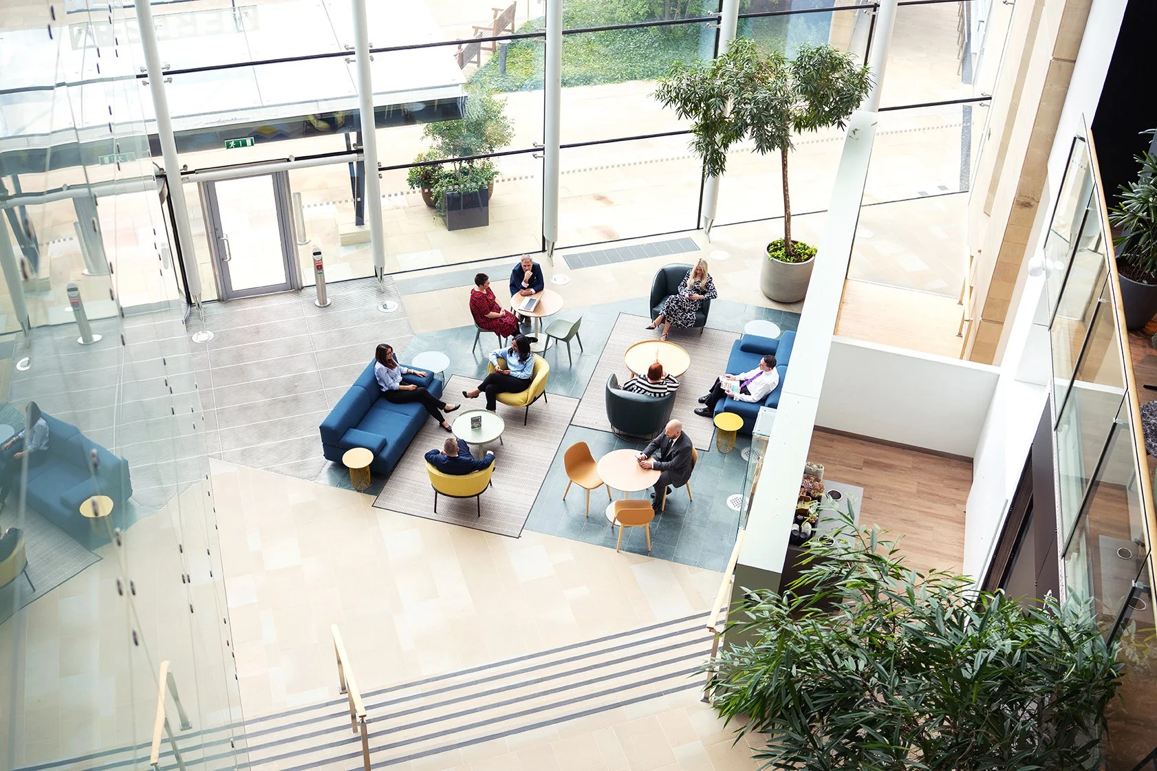 Interior of a modern office or lobby with large glass windows. Several people are sitting and talking on sofas and chairs arranged in small groups, with some using laptops. There are potted plants and a glass railing visible in the space.
