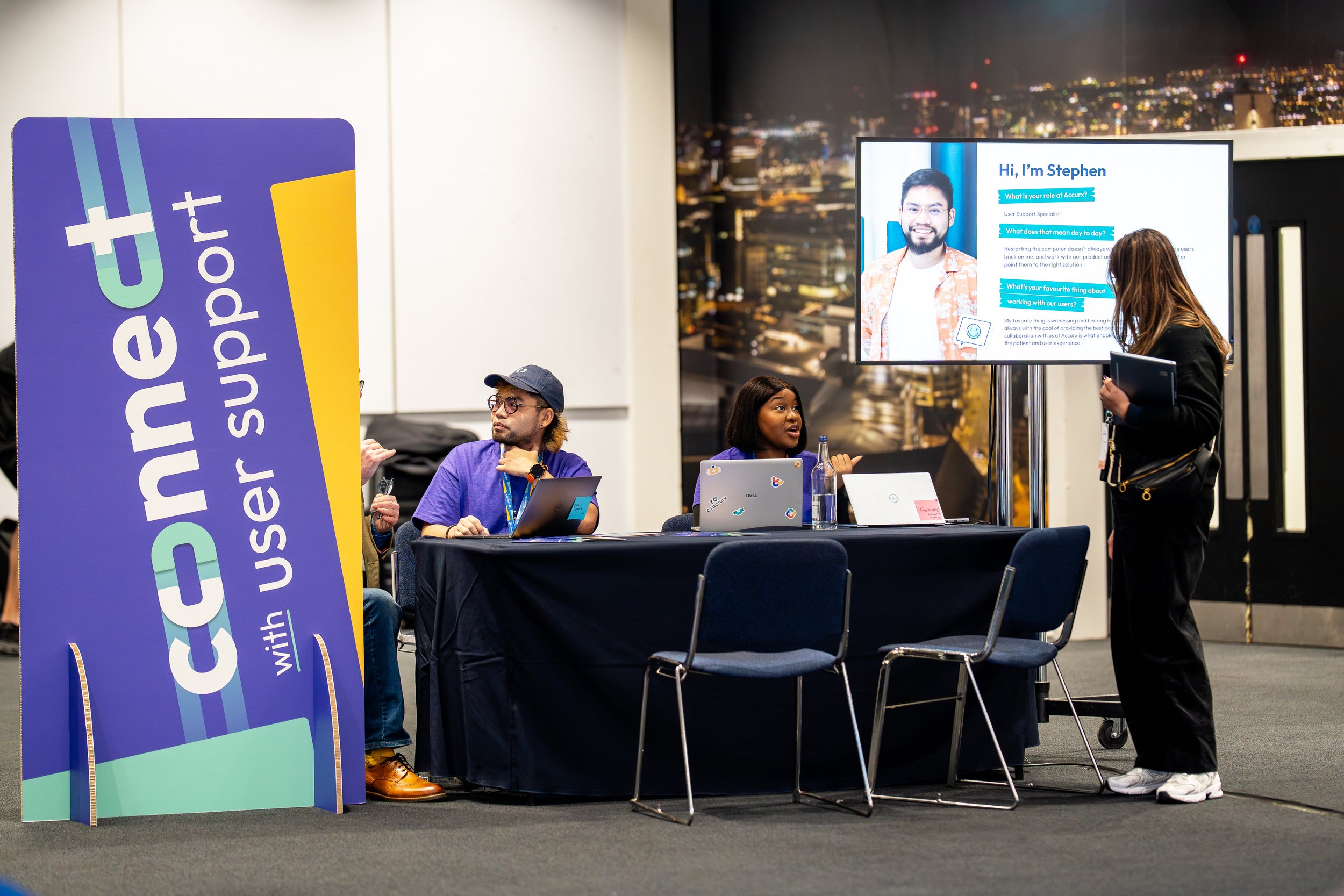 At a conference booth with a large vertical sign that reads 'Cure with support,' three people are engaged in a discussion. One person is sitting at a table with a laptop, another is standing and speaking, and the third is sitting with a laptop in fro