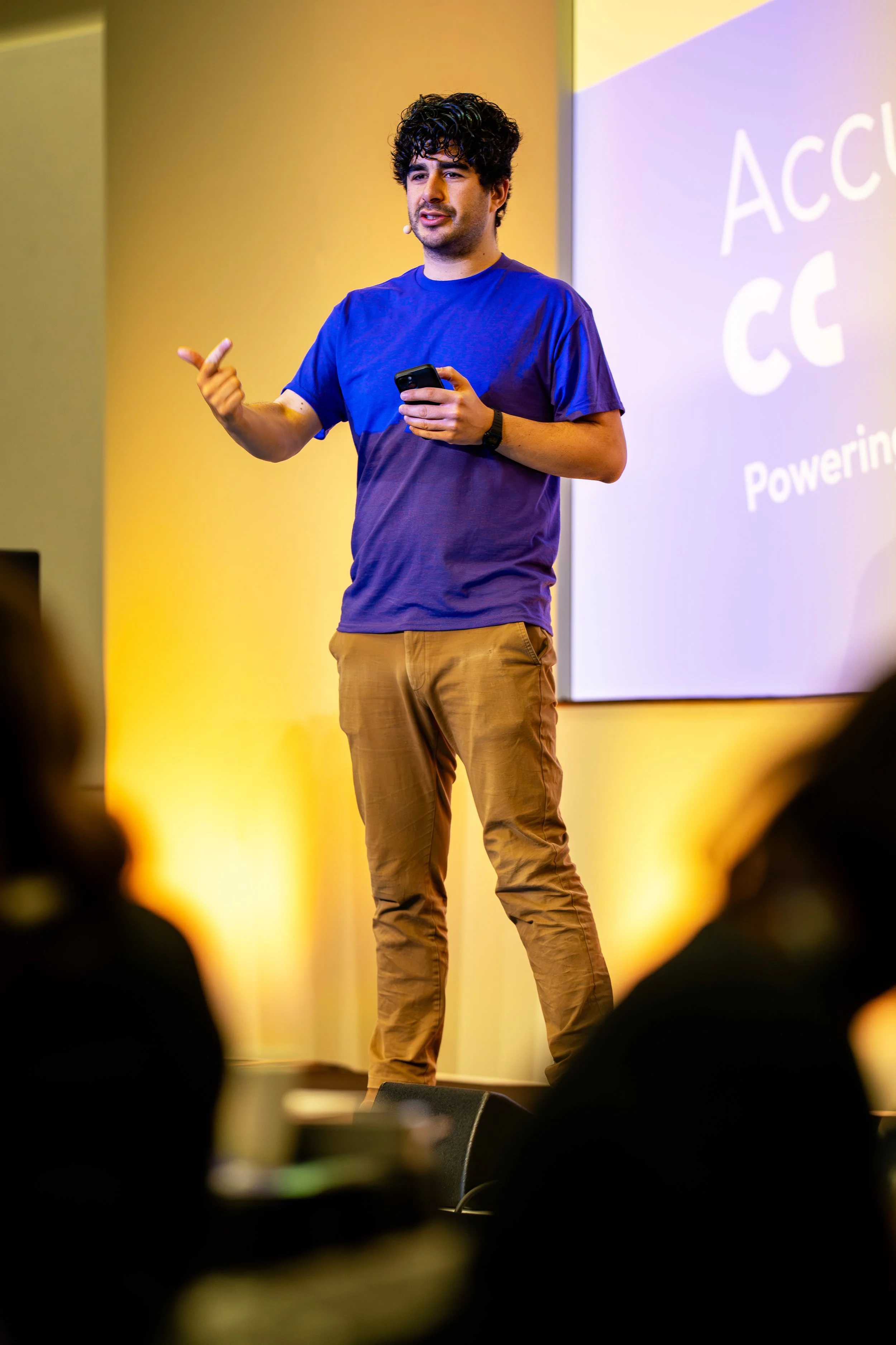 Young man with curly dark hair, wearing a blue T-shirt and tan pants, speaking or presenting on stage with a microphone headset, holding a smartphone, in front of a projection screen with partial text.