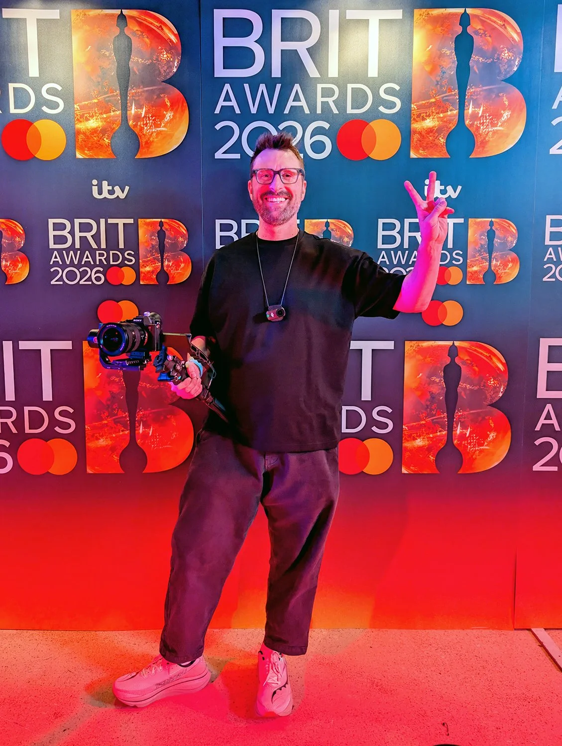 A man smiling and making a peace sign with his right hand, holding a camera stabilizer with a camera attached in his left hand at the 2026 BRIT Awards, standing in front of a branded backdrop.