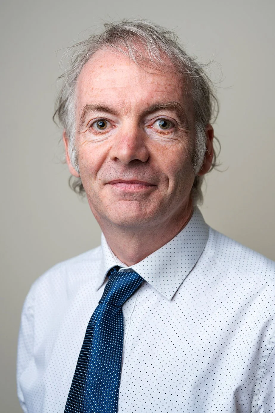 A front-facing portrait of a middle-aged man with light-colored hair, blue eyes, wearing a white dress shirt with small dark dots and a dark blue tie, standing against a plain light-colored background.