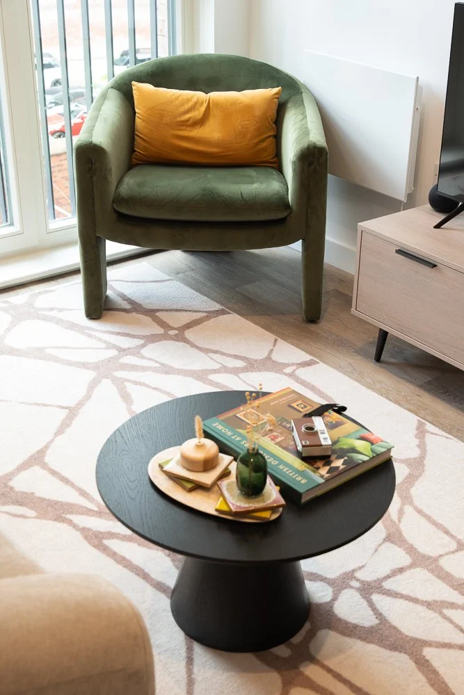 A green armchair with a yellow pillow, a round black coffee table with books, candles, and decorative items, a light-colored area rug, and a flat-screen TV on a light wood media stand in a bright living room.