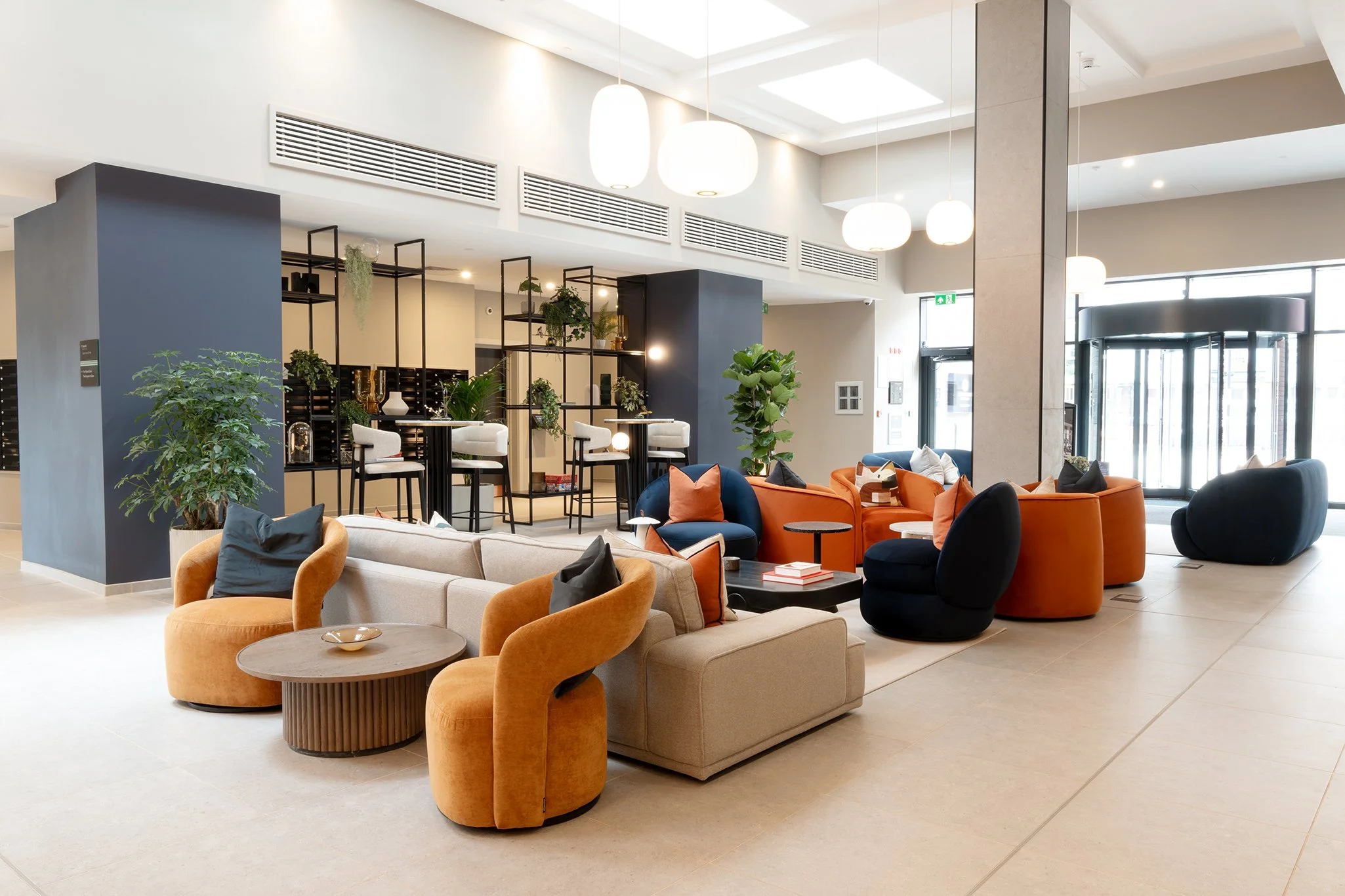 Modern hotel lobby with seating area including beige, blue, and orange armchairs, and tables, decorated with plants, pendant lights, and large windows.