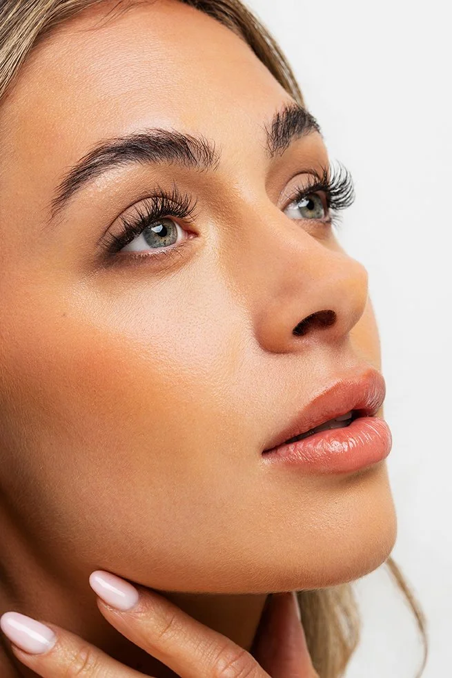 Close-up of a woman with flawless skin, blue eyes, and light makeup, touching her chin gently.