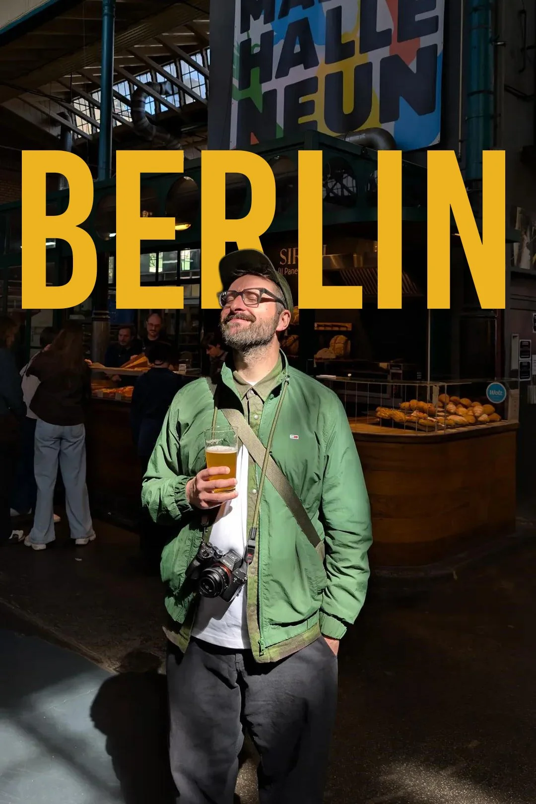 A man in a green jacket holding a glass of beer, standing inside a market or food hall in Berlin, with a camera hanging around his neck and people in the background near a bakery stall.