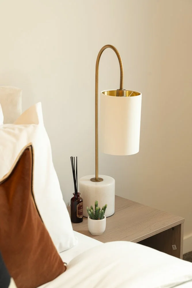 A bedside table with a modern lamp, plant, and reed diffuser next to a bed with white bedding and a brown leather pillow.