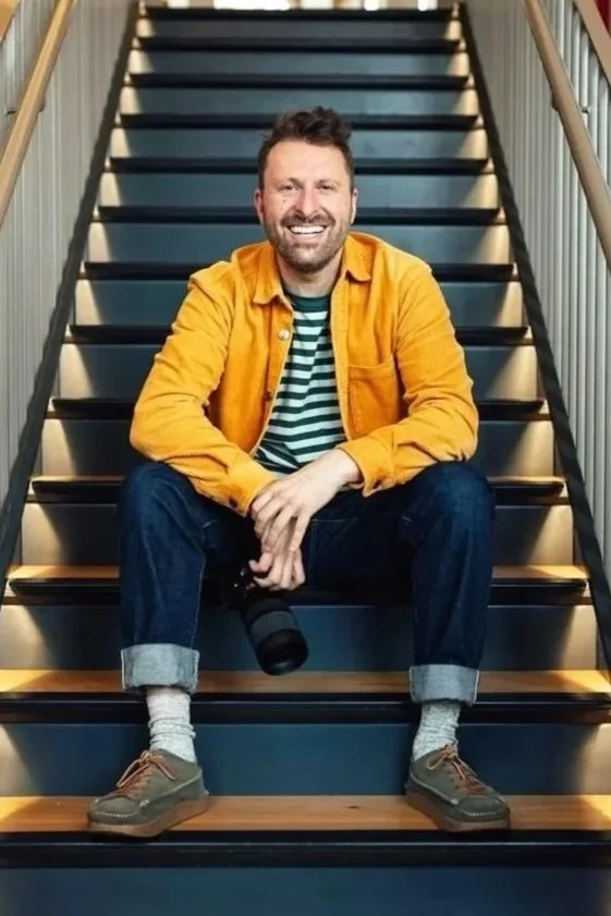 A smiling man sitting on stairs, wearing a yellow jacket, striped shirt, jeans, and brown shoes, holding a camera.