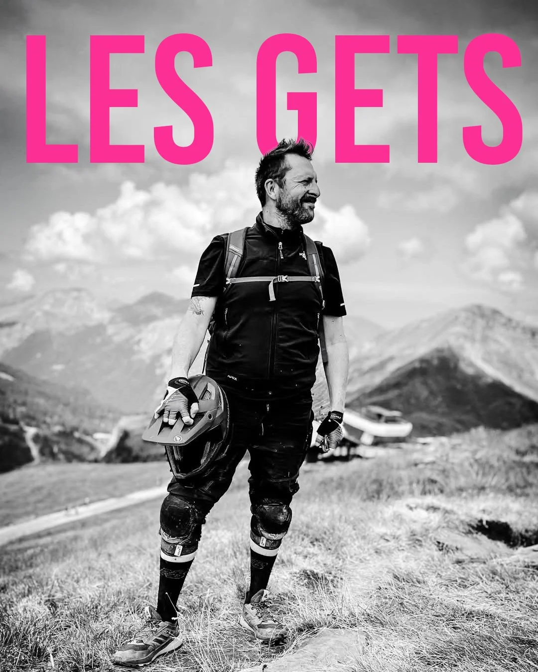 Black and white outdoor photo of a man in mountain biking gear standing on a grassy trail with mountains and clouds in the background, holding a helmet, with the text 'LES GETS' in bold pink letters at the top.