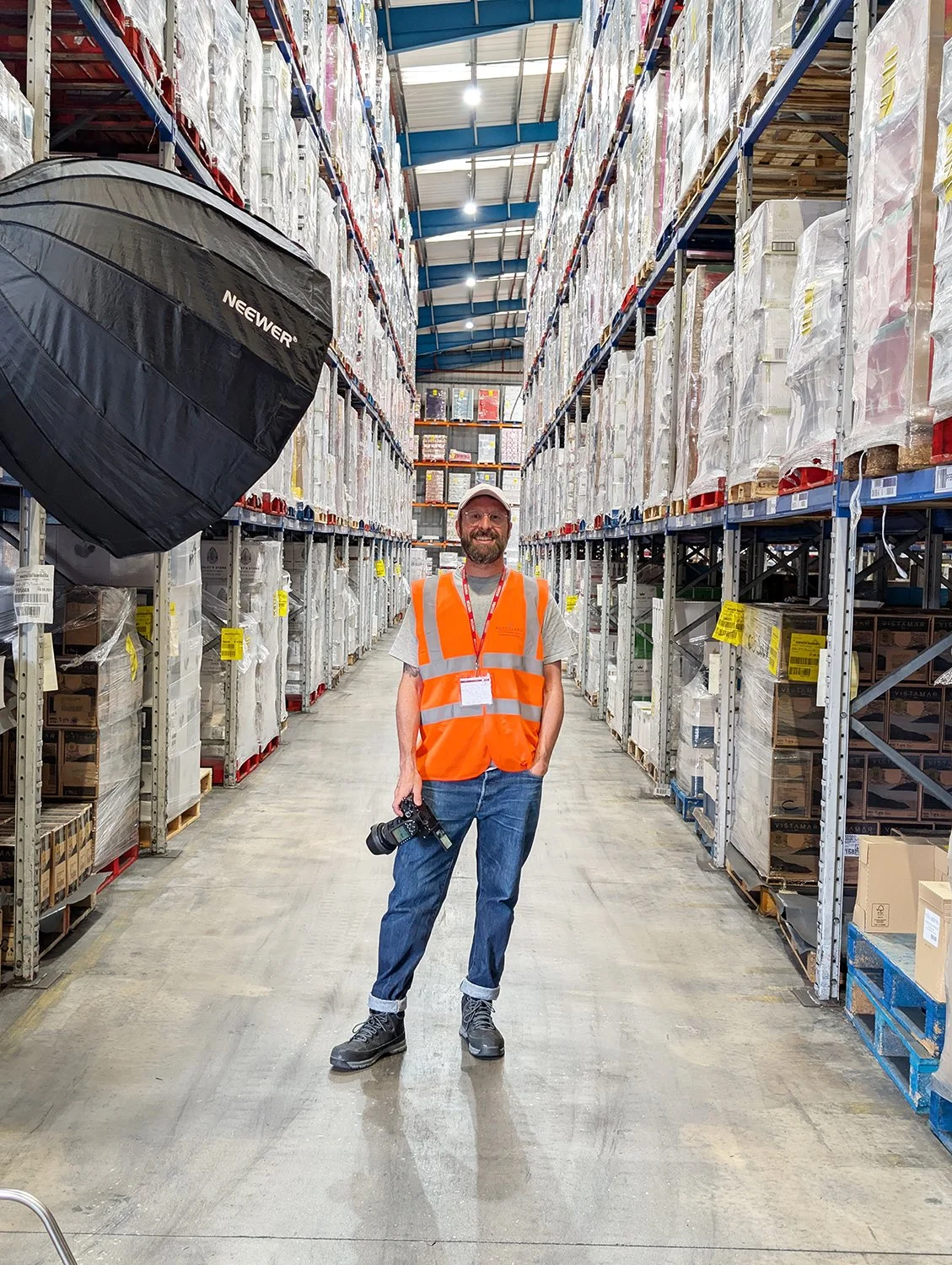 A man wearing an orange safety vest, gray t-shirt, jeans, and boots, smiling and holding a camera, standing in a warehouse aisle with tall shelves stocked with boxed goods.
