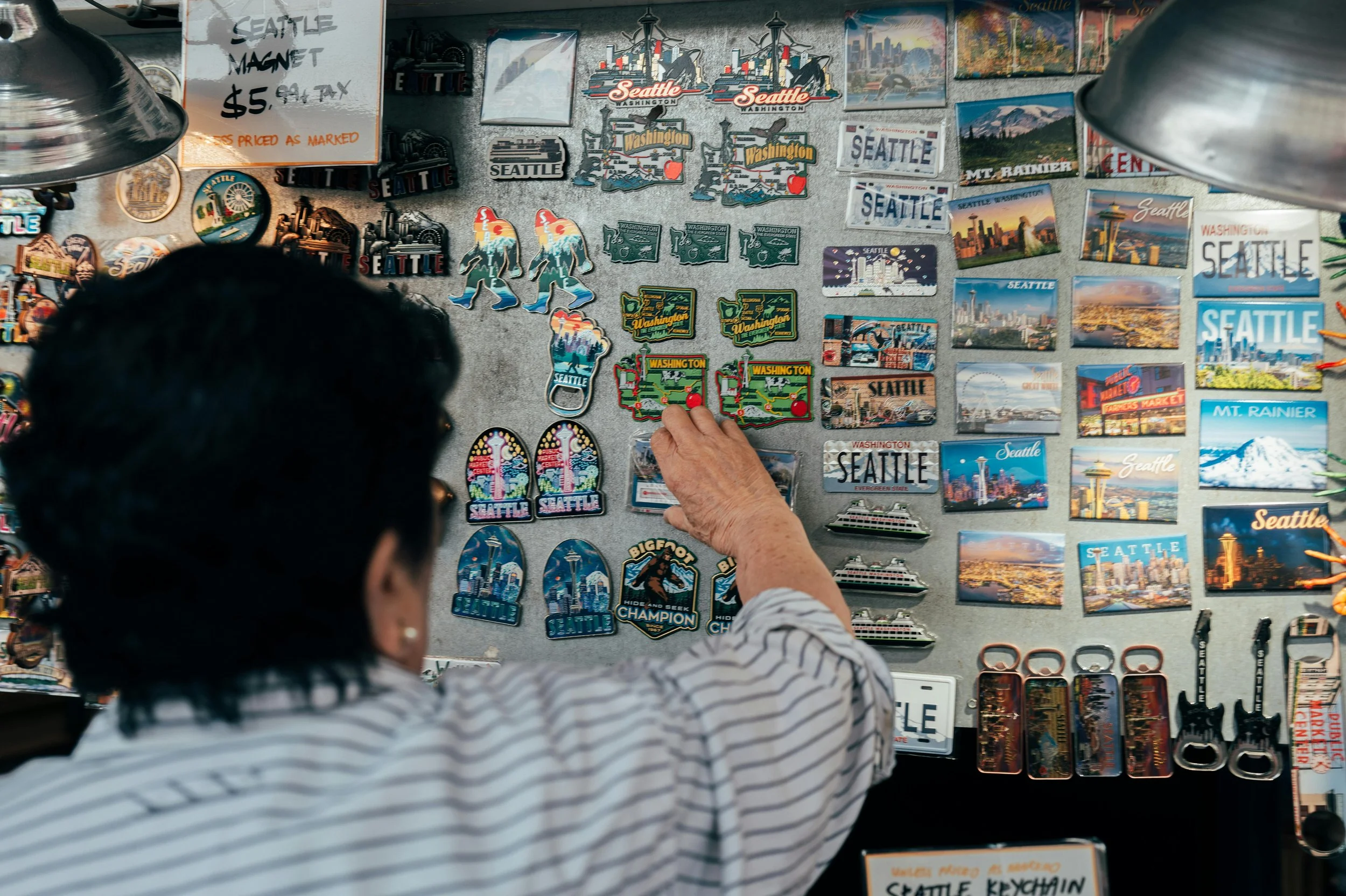 People browsing a stall with various Seattle-themed souvenirs, magnets, and postcards in a market setting.