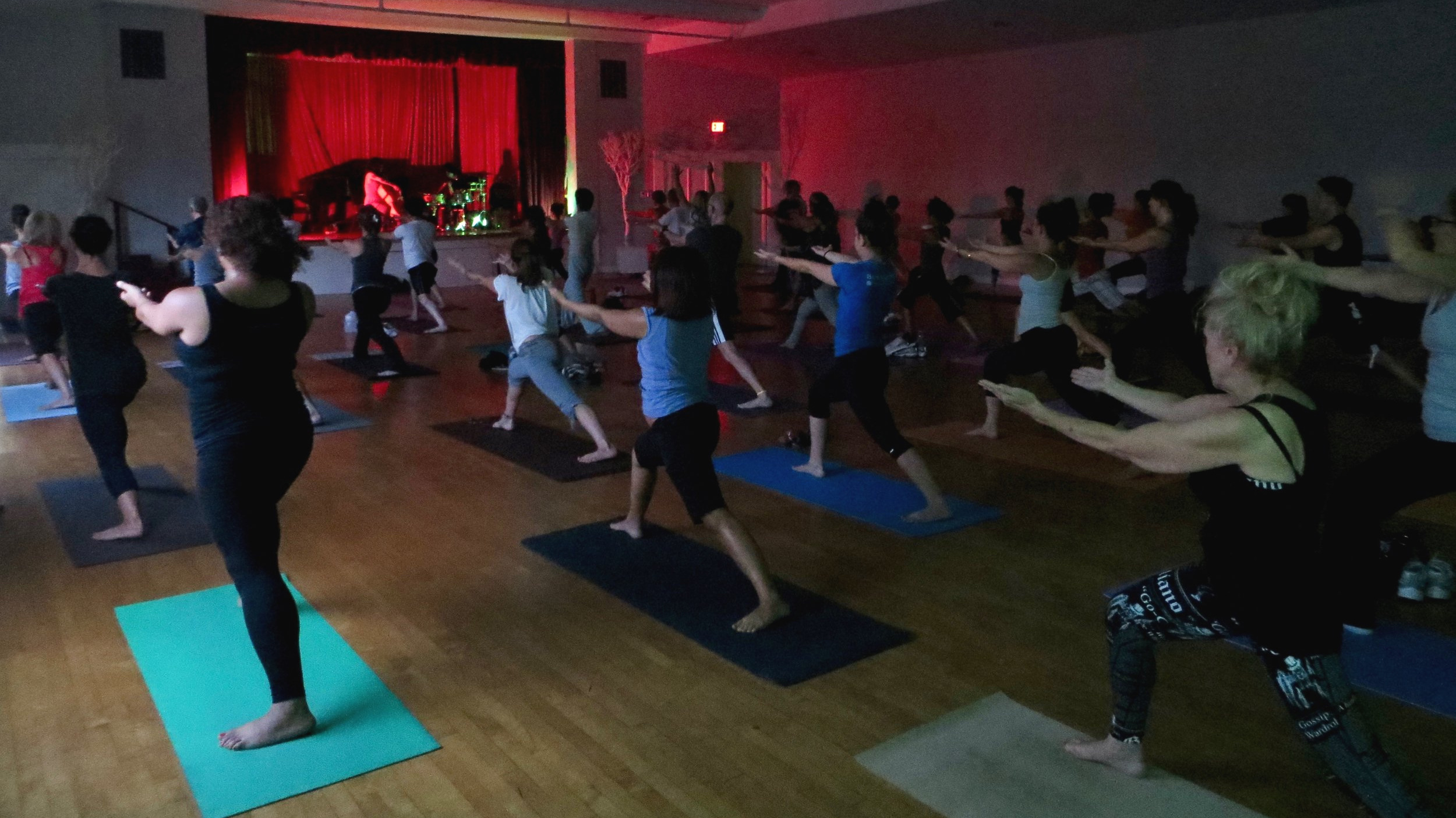 Black Market Yoga class performing a variation of warrior one pose in a large auditorium with low lights and a musical guest at the piano.jpg