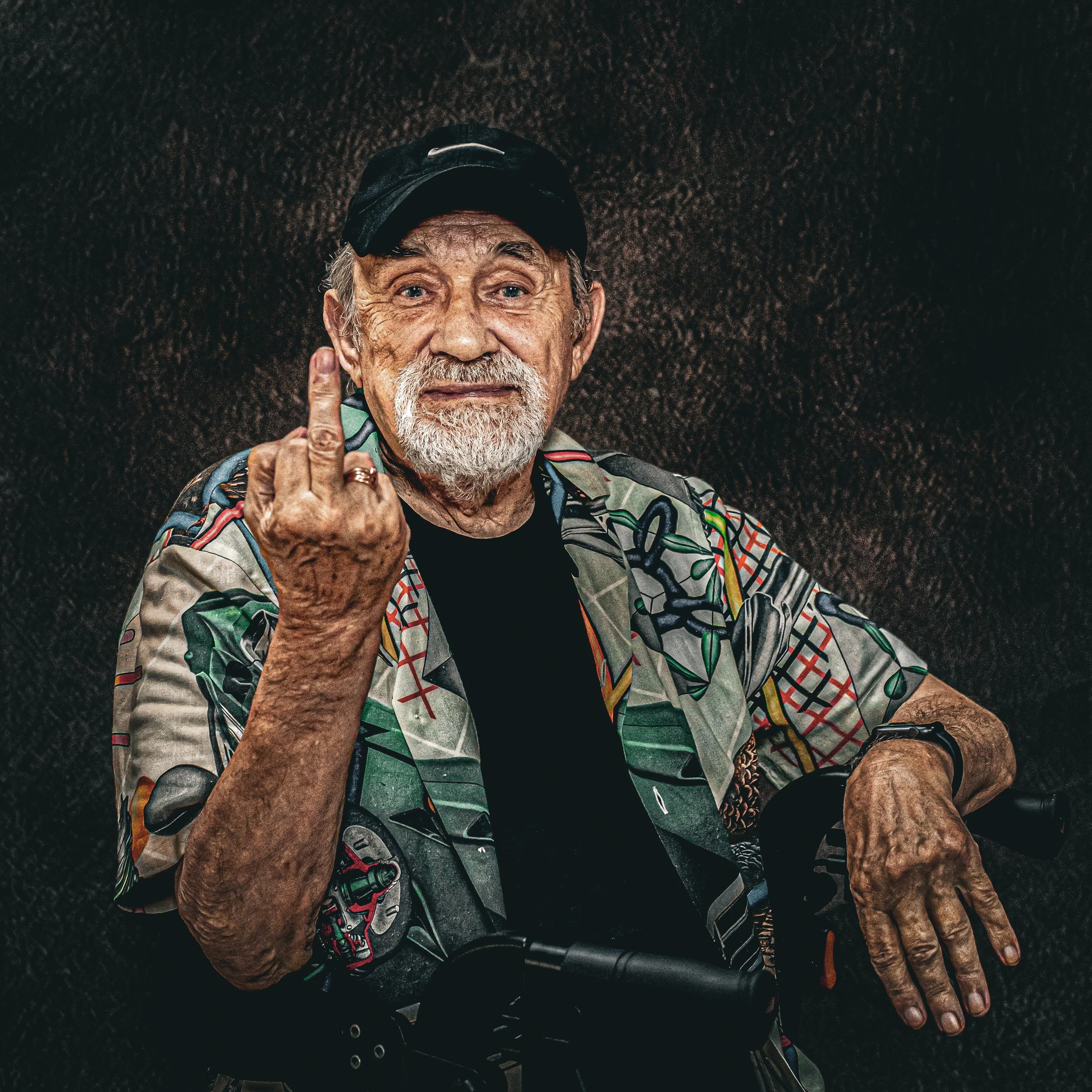 An elderly man with a white beard and mustache wearing a colorful patterned shirt and a black cap, making a rude gesture with his middle finger.