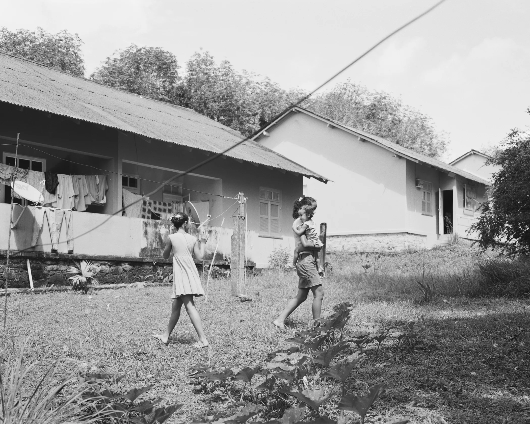 Sisters Ivna and Ingrid Souza playing with cousin Monick Farias in the family backyard with squash plants in the foreground at company housing complex Vila 4, April, 2014, Michelin Rubber Plantation, Bahia, Brazil. 2014/2018