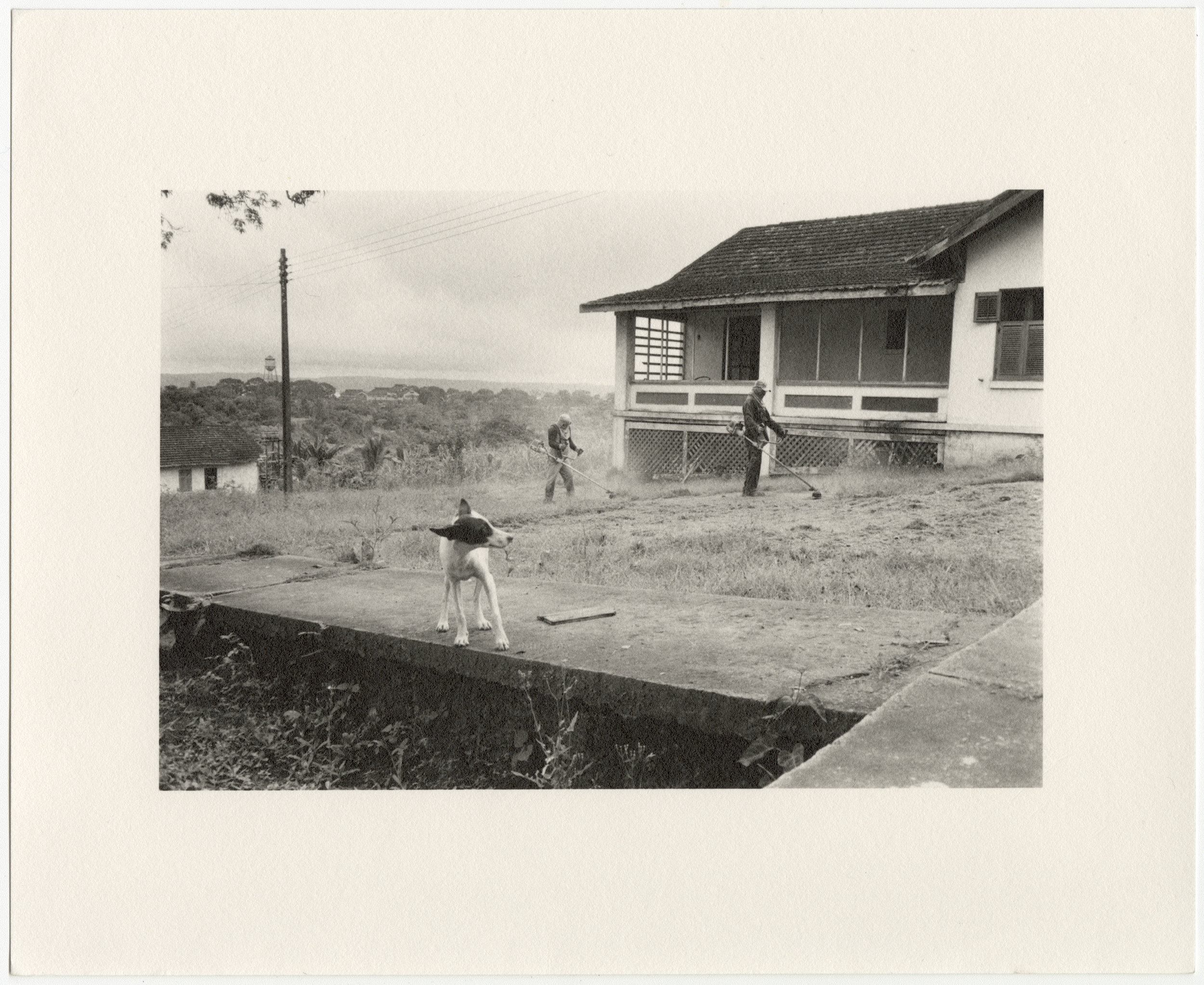  Fordlândia manager’s house on Vila Americana, with men weedwacking the front yard and dog, built in 1929 by the Companhia Ford Industrial Do Brasil Ford Motor Company. 2014, Fordlândia, Pará, Brazil. Gelatin silver fiber print, 8” x 10”, 2014/2018 