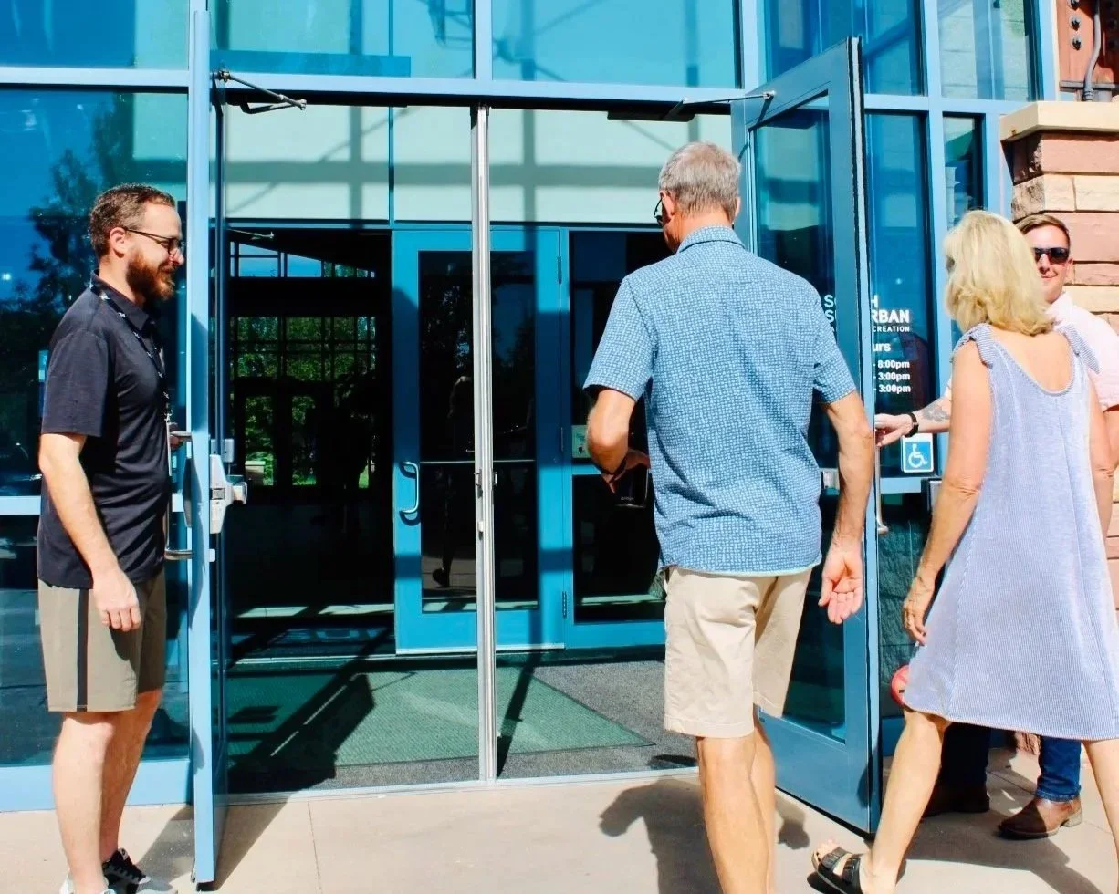 A group of four people standing outside the entrance of a building, with some individuals holding the door open. The building has a glass facade, and the door has a wheelchair accessibility sign.