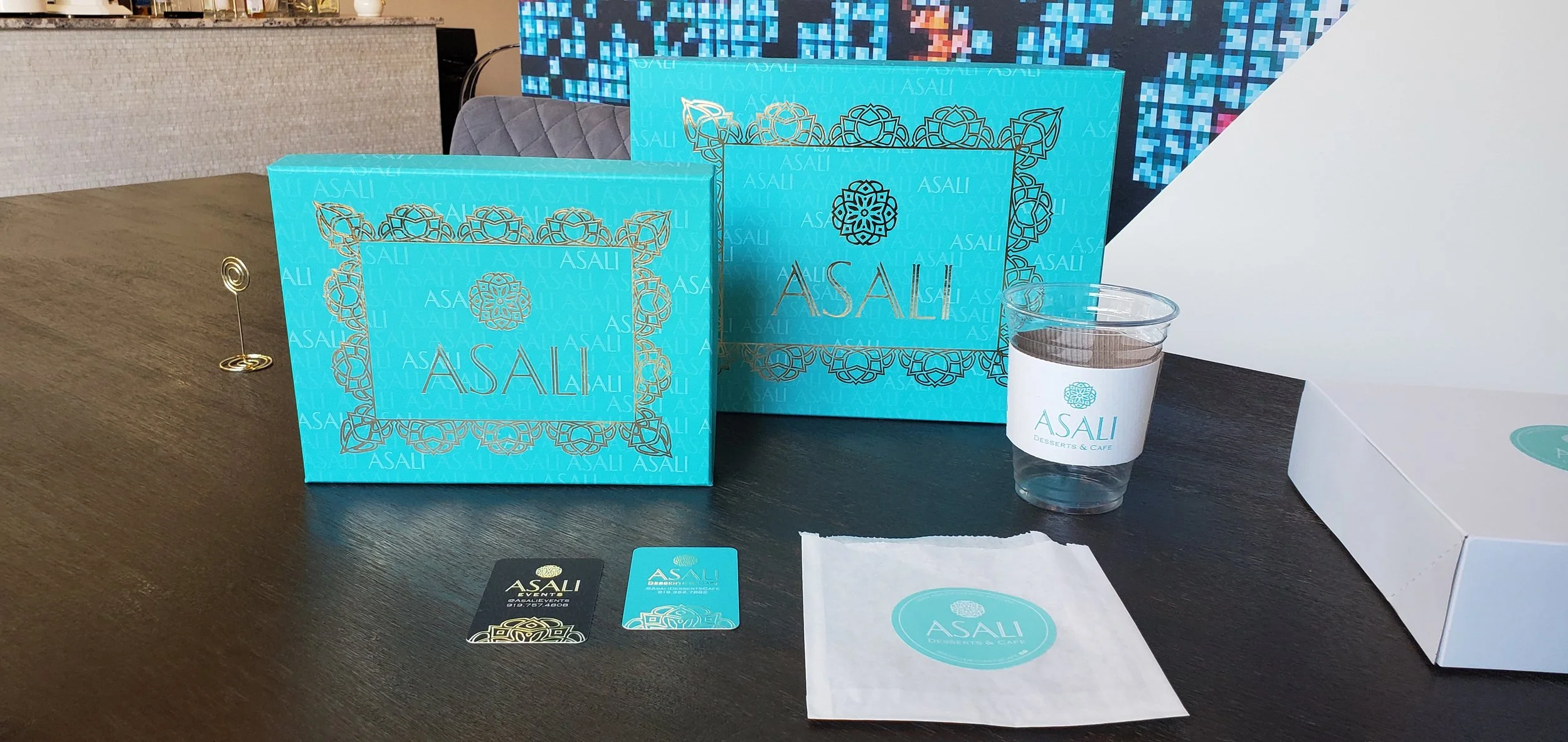 Blue ASALI gift boxes, a printed cup with ASALI logo, business card, and napkin with ASALI logo on a dark table.
