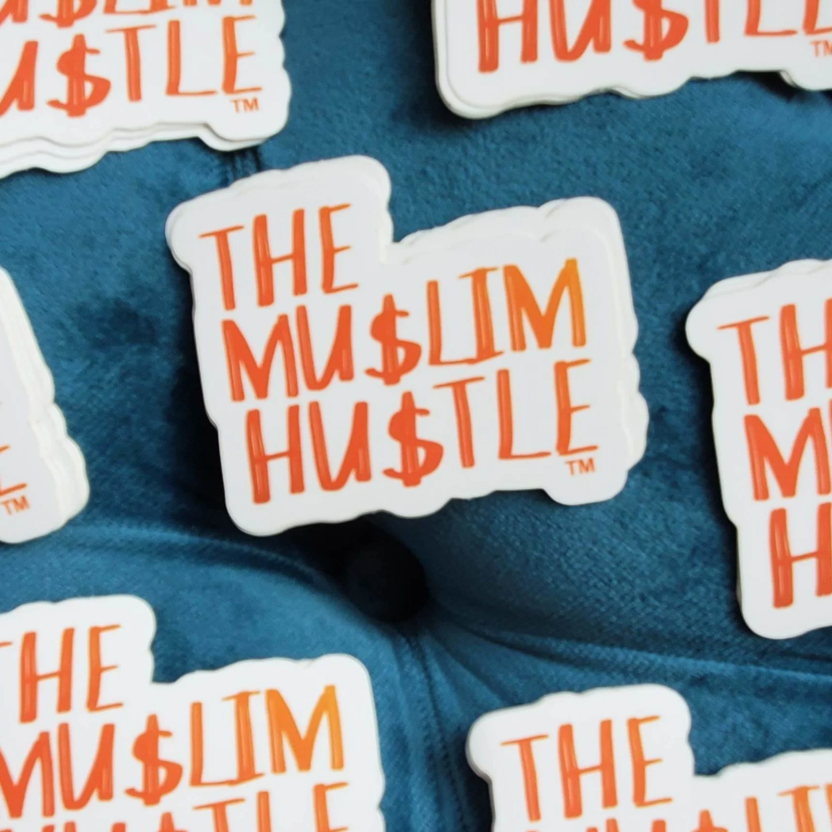Stickers with the phrase "The Muslim Hustle" in orange text on a white background, displayed on a blue surface.
