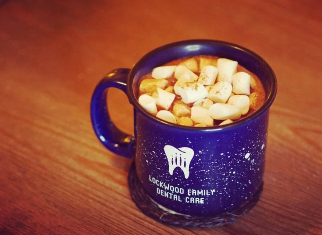 Coffee mug with marshmallows on top, featuring the logo and name of Lockwood Family Dental Care, placed on a wooden surface.