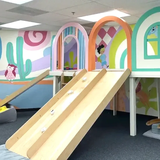 Indoor play area featuring a wooden ramp with climbing holds, colorful arches, and wall murals with cartoon characters and abstract designs.