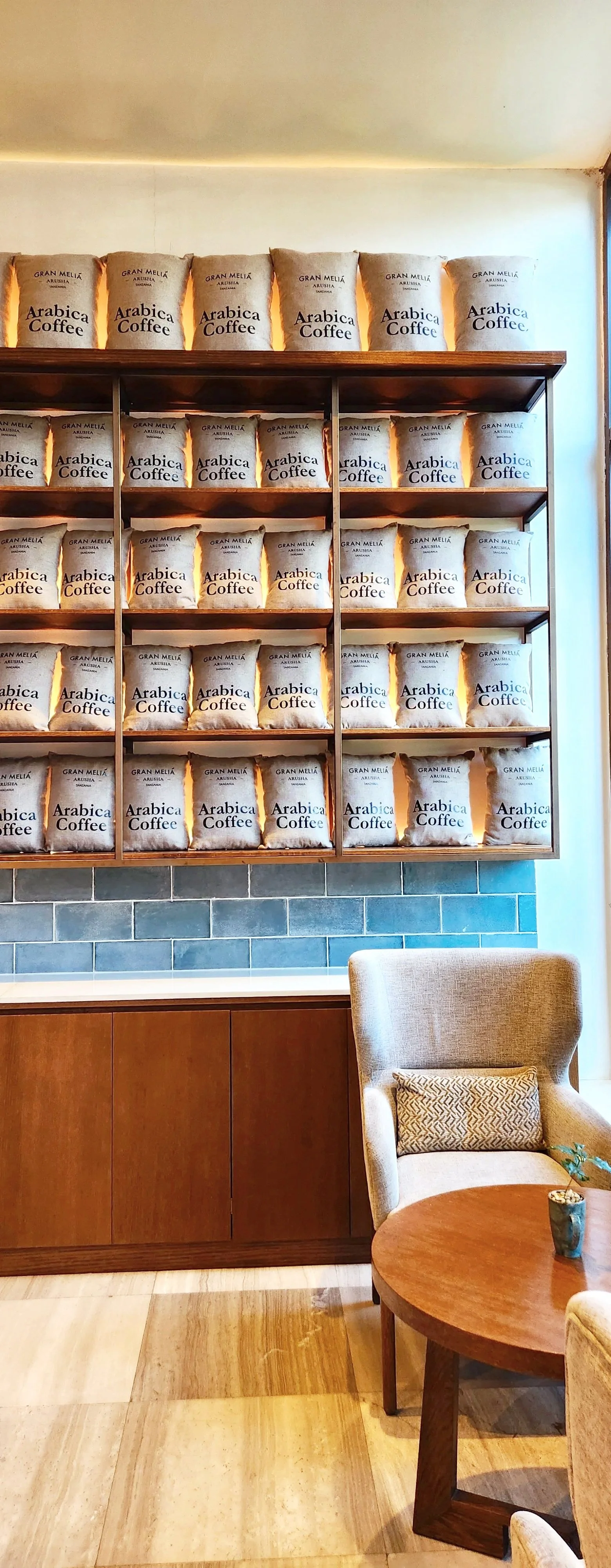 A wall display of coffee bags labeled 'Arabica Coffee' and 'Gran Melia Arusha, Tanzania' in a cozy cafe setting with a beige armchair, round wooden table, and a small potted plant.