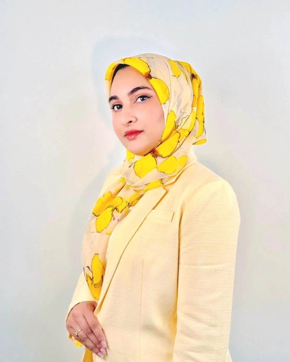 Yumna Ali. A woman wearing a yellow blazer and a floral yellow and beige hijab standing against a plain light-colored background.