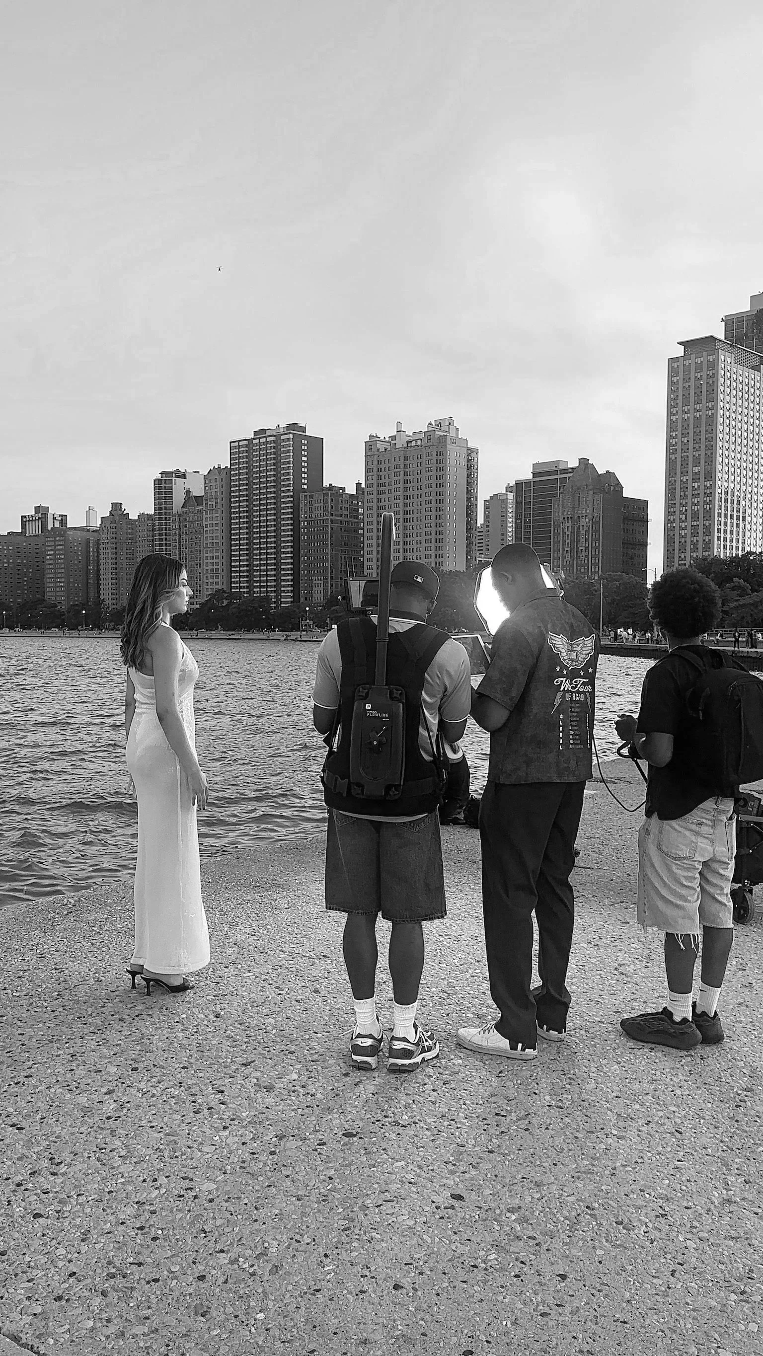 A woman in a white dress is standing near a river with city buildings in the background. Three men with filming equipment are filming her, with the sun reflecting off their equipment.