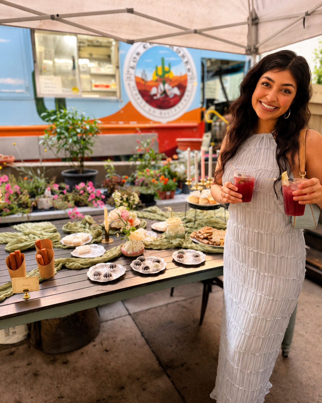 A woman in a white dress smiling and holding two red cocktails at an outdoor market stall with desserts and flowers.